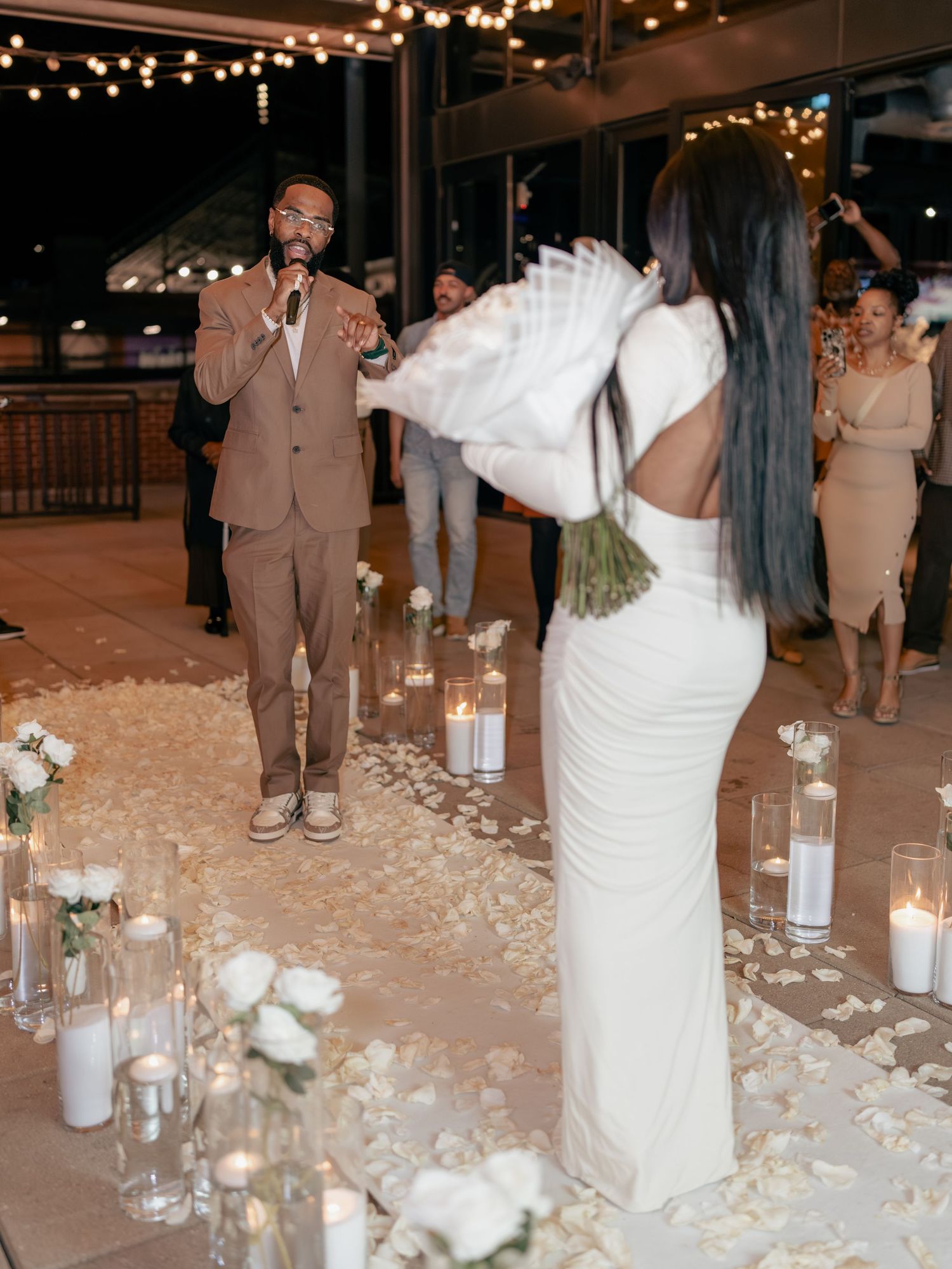 Elegant outdoor wedding ceremony at night with candles, rose petals, and string lights as bride holds white fan bouquet.