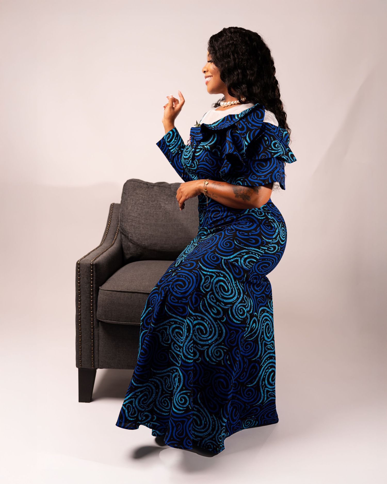 Woman in blue and black patterned dress poses elegantly with grey armchair in studio setting.