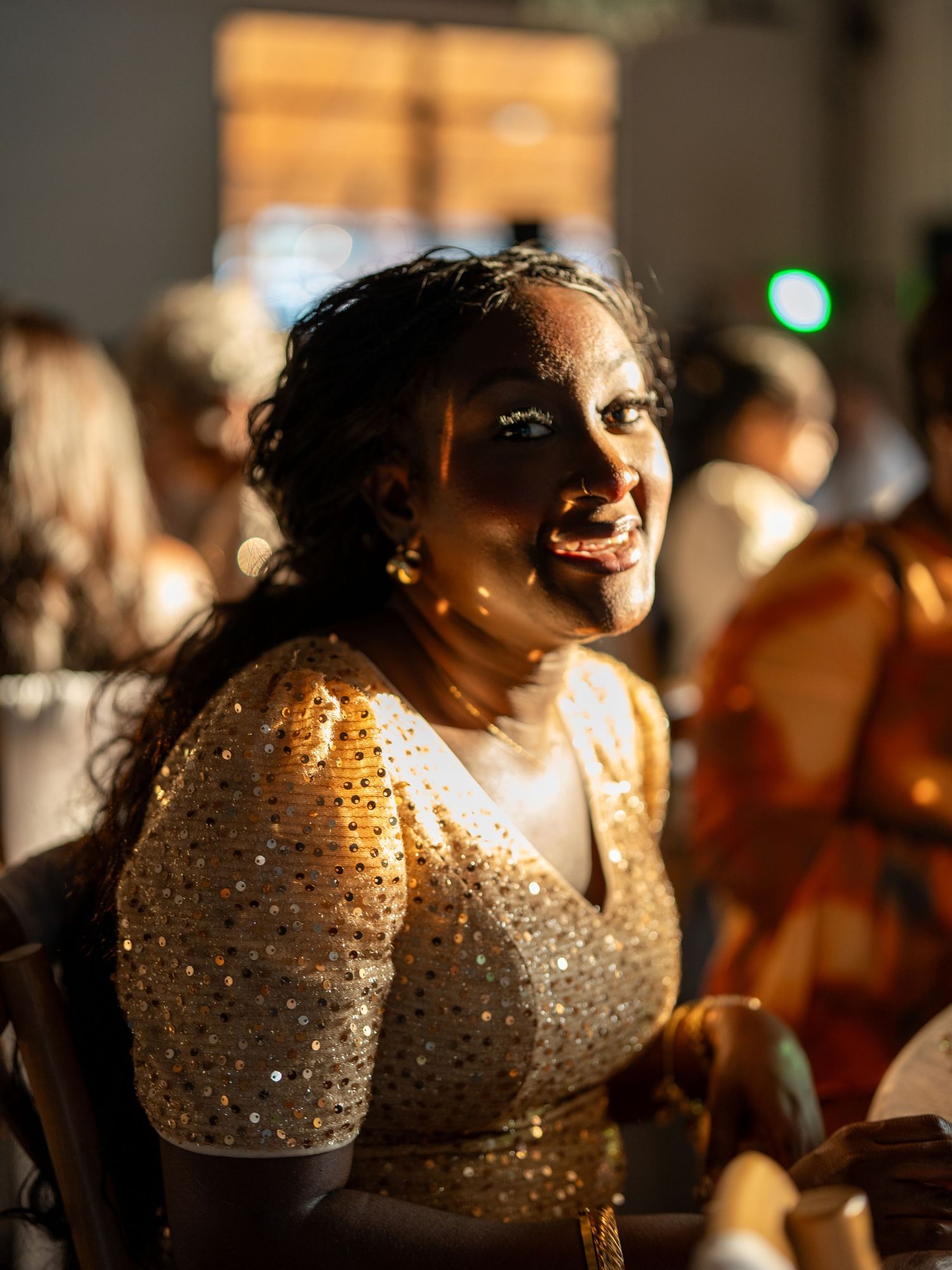 A smiling woman in a gold sequined dress enjoys a festive event, bathed in warm golden light.