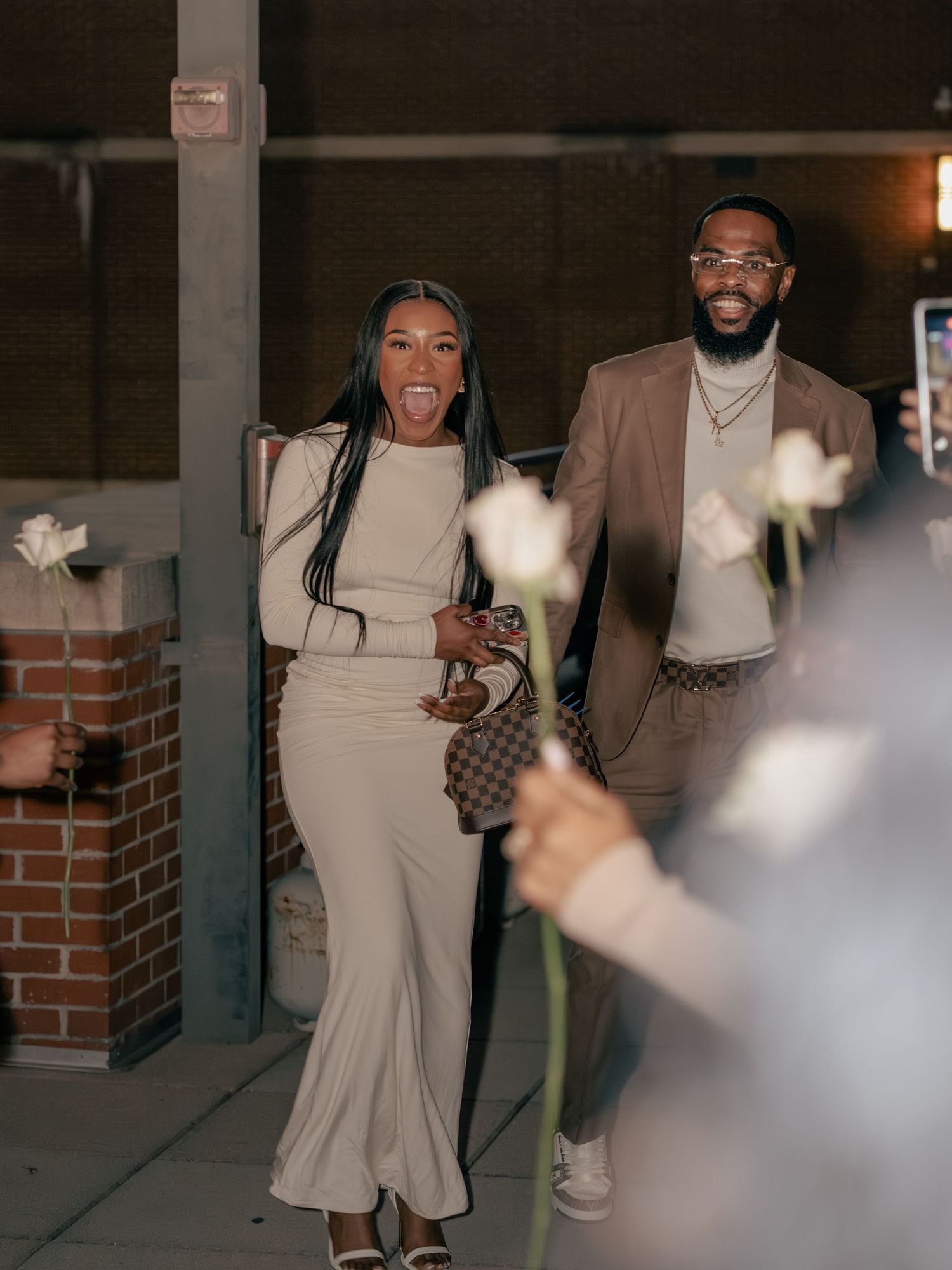 Couple in coordinating beige outfits smiling outdoors at night, woman in maxi dress with Louis Vuitton bag, man in blazer.