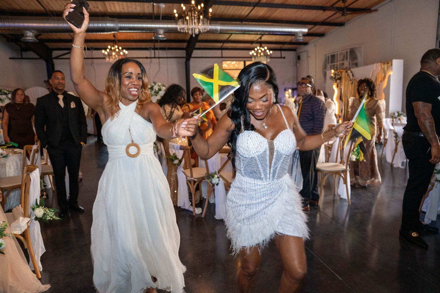 Two women dancing joyfully at a wedding reception, one in white bridal gown, other holding Jamaican flag in festive venue.