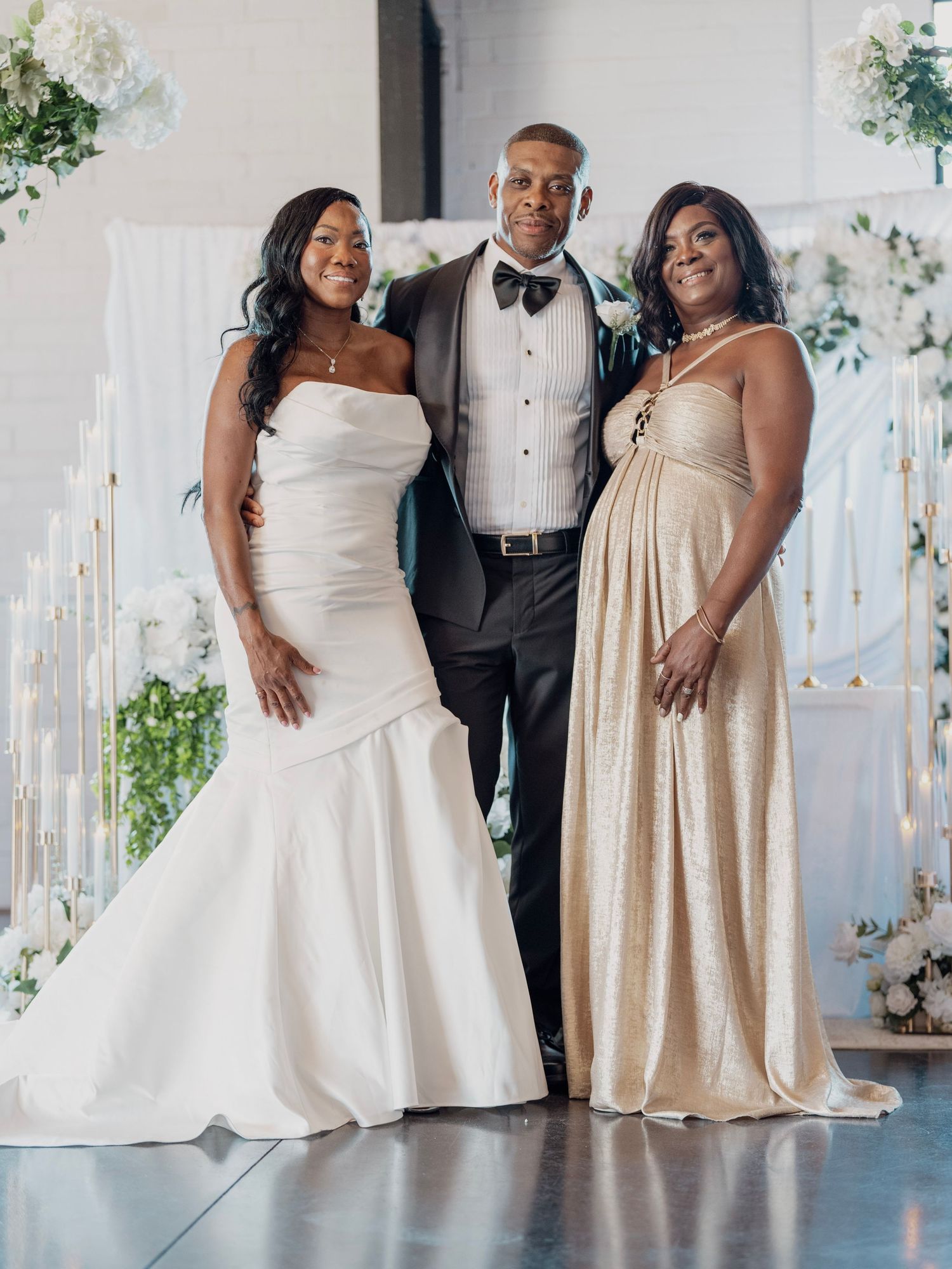 Bride in a white strapless gown stands with a man in a tuxedo and bow tie and a woman in a gold gown at a wedding with white floral decor.