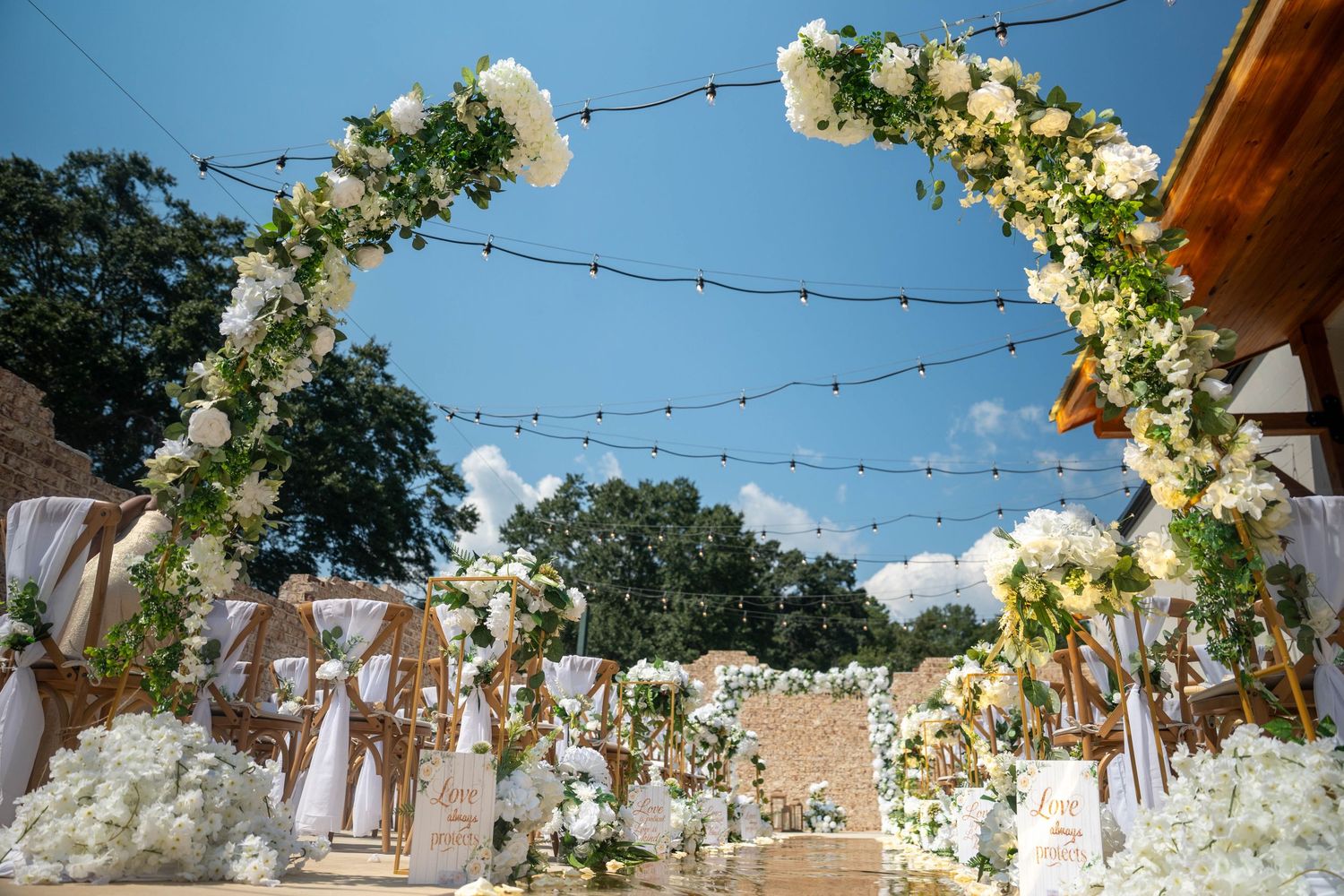 Elegant outdoor wedding ceremony arch adorned with white roses and greenery under string lights and blue sky.