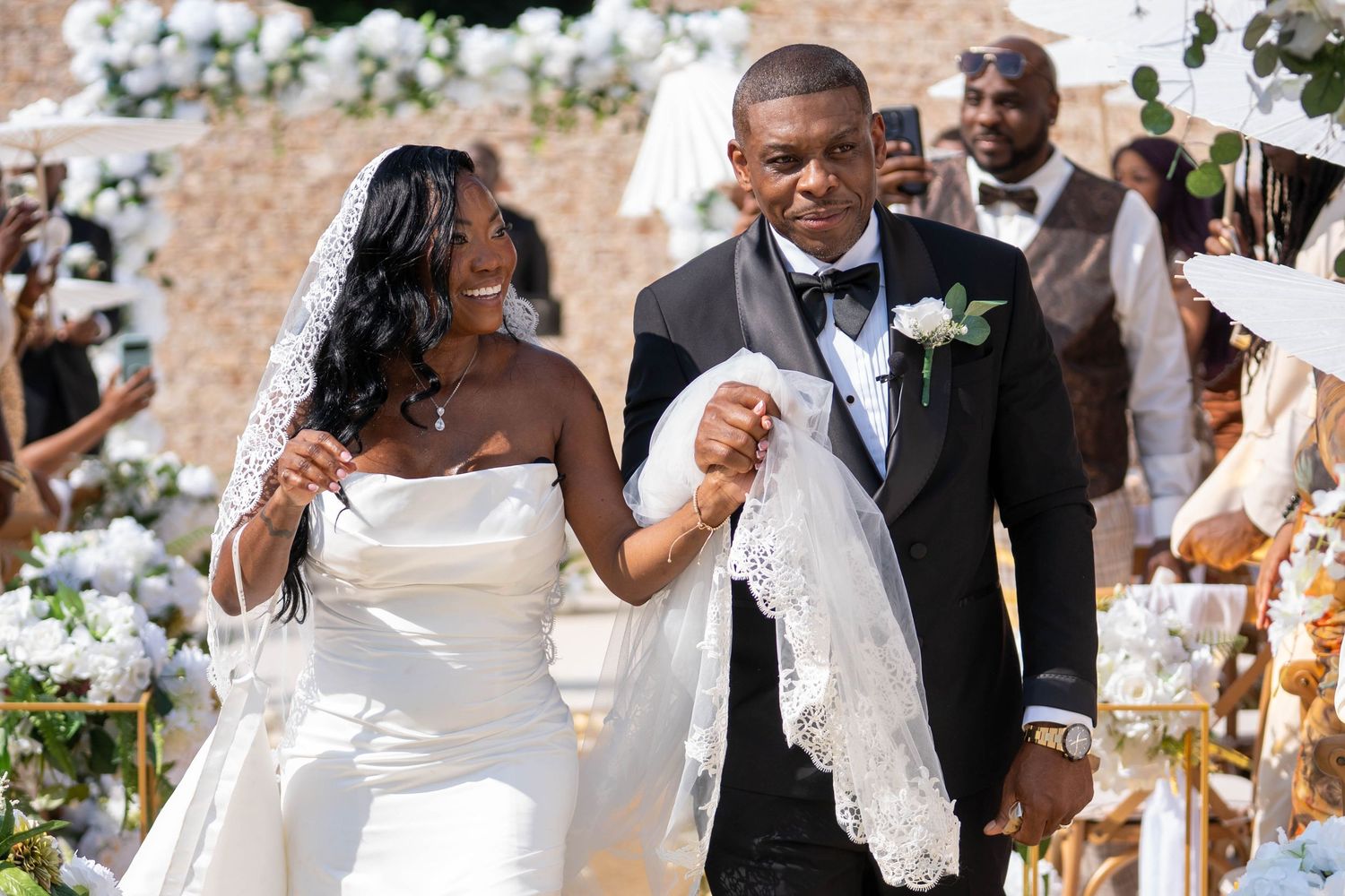 A joyful bride and groom walk down the aisle at an elegant outdoor wedding ceremony with white floral decor.
