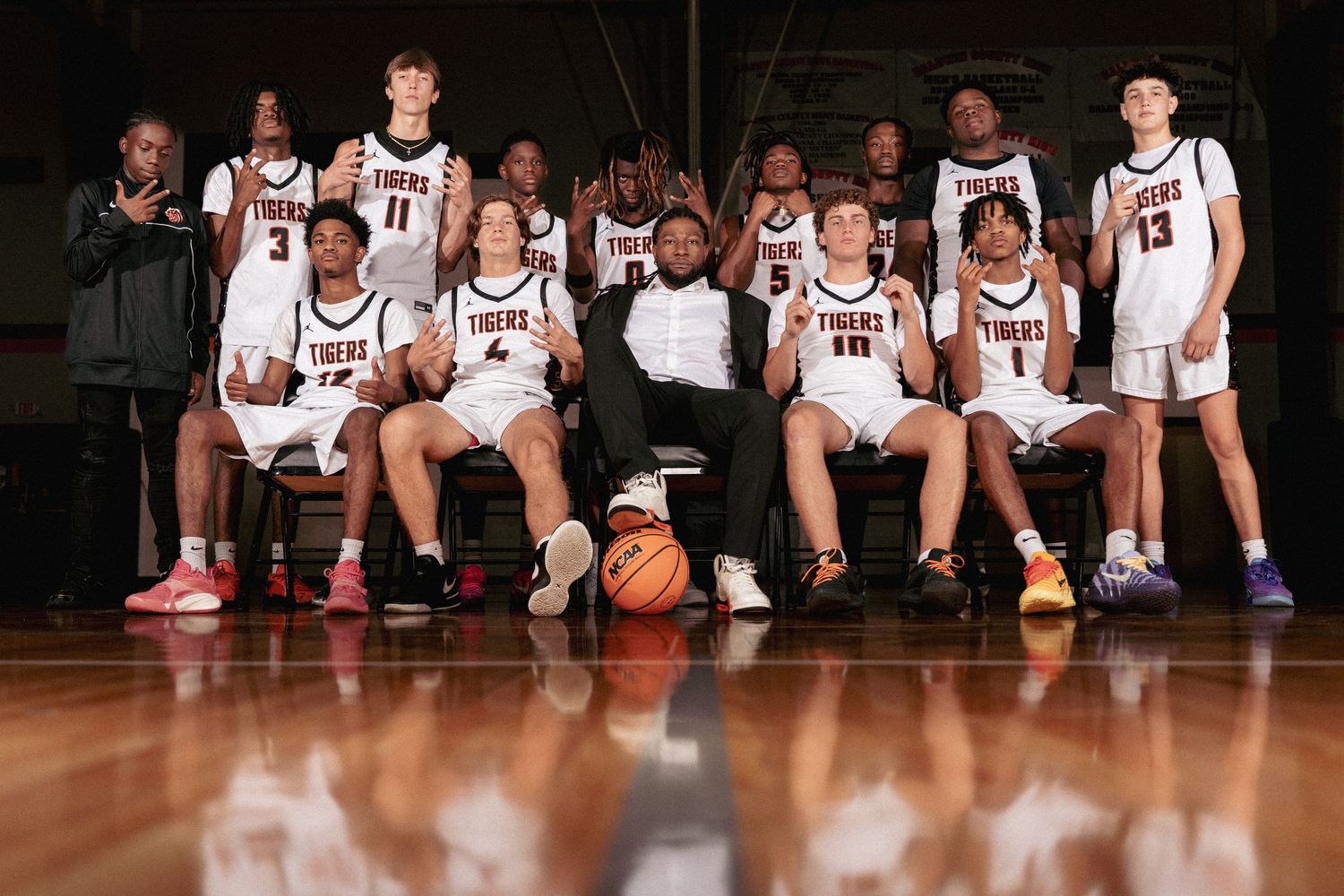 Basketball team sits on bench in white uniforms while posing for a panoramic photograph in gymnasium.