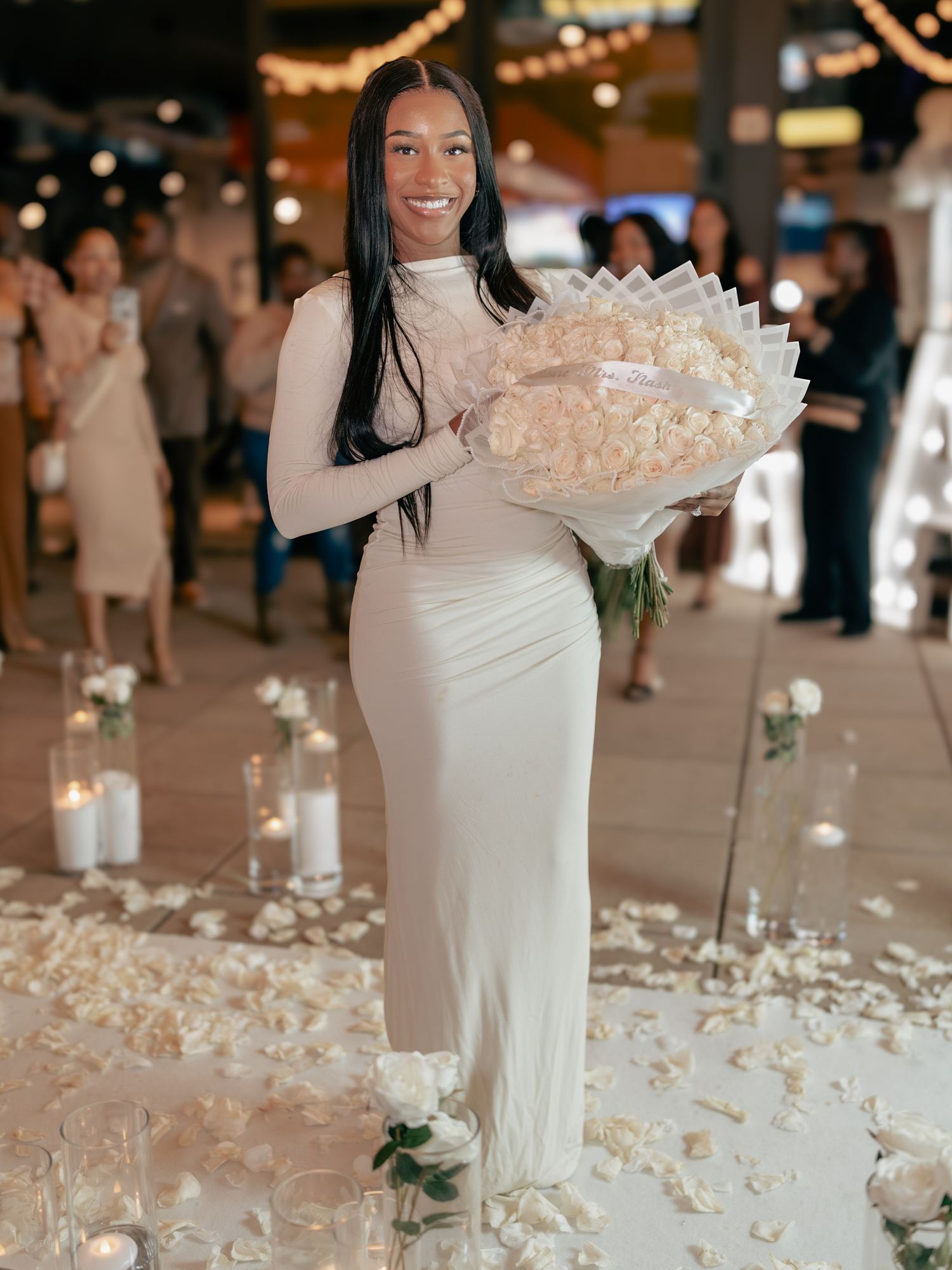 Woman in white dress holding large bouquet of white roses stands among candles and rose petals at elegant event.