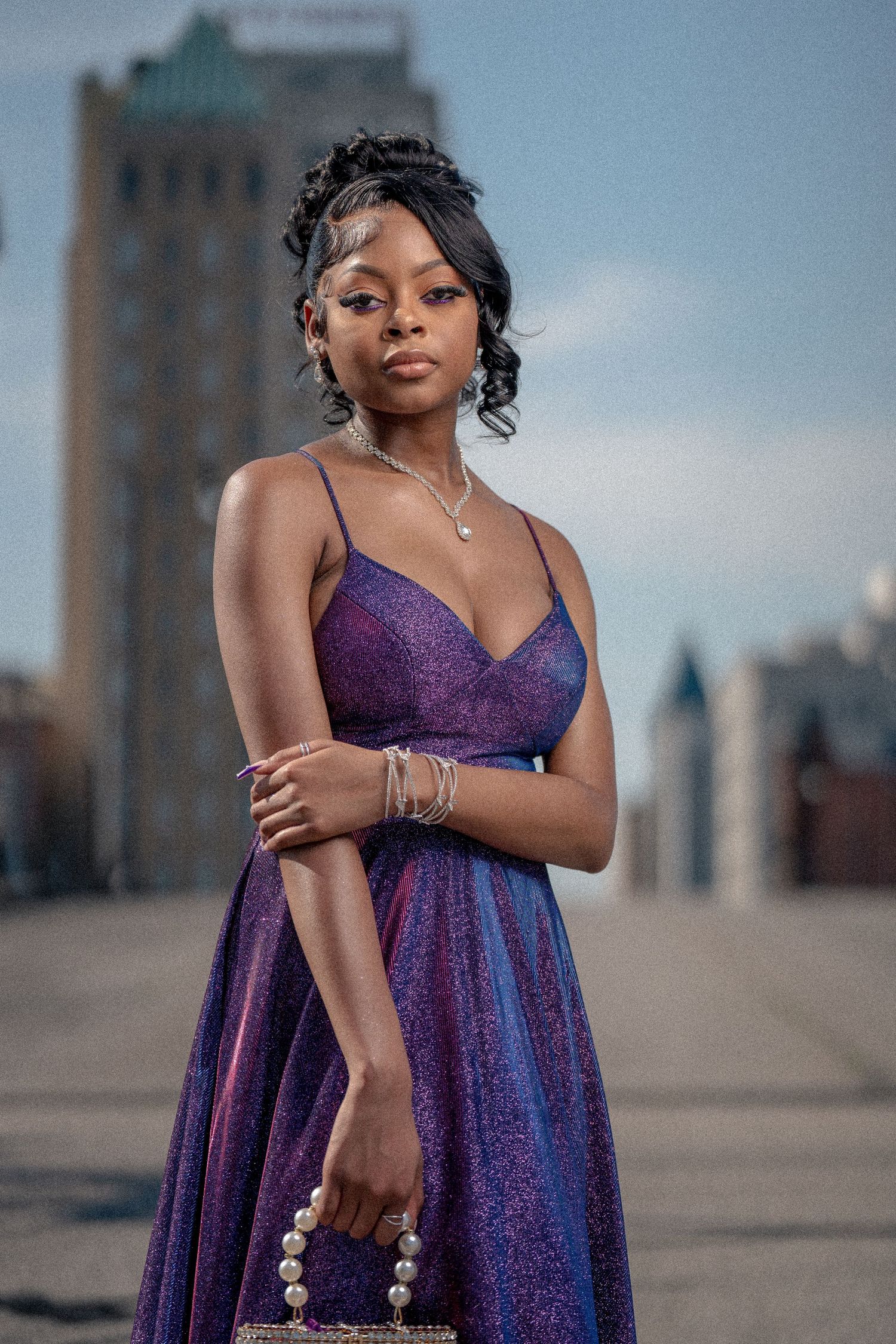 Portrait in a shimmering purple evening gown with delicate jewelry standing in an urban setting at dusk.