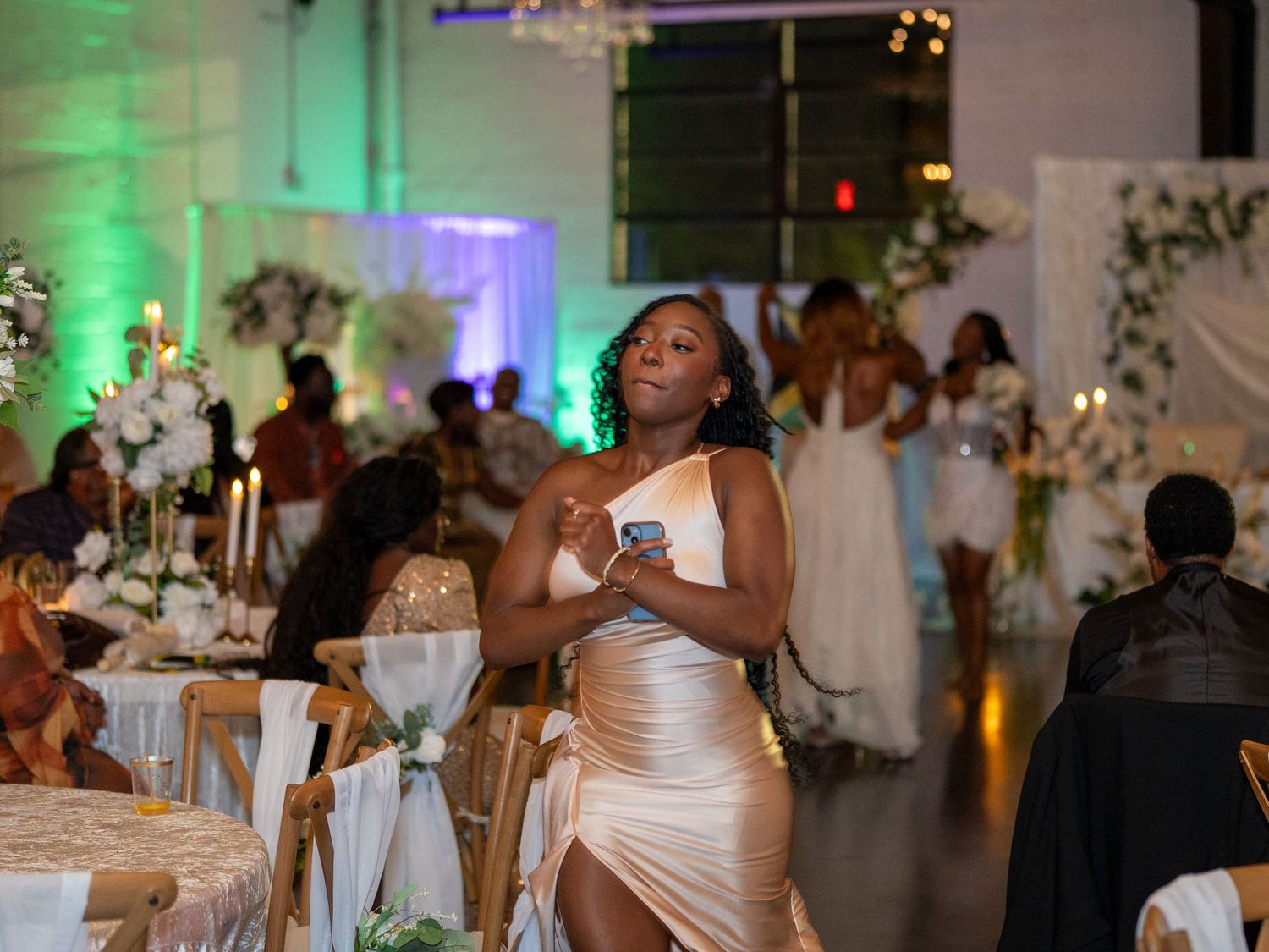 Woman in elegant champagne satin dress dancing at a formal wedding reception with decorated tables and floral arrangements.