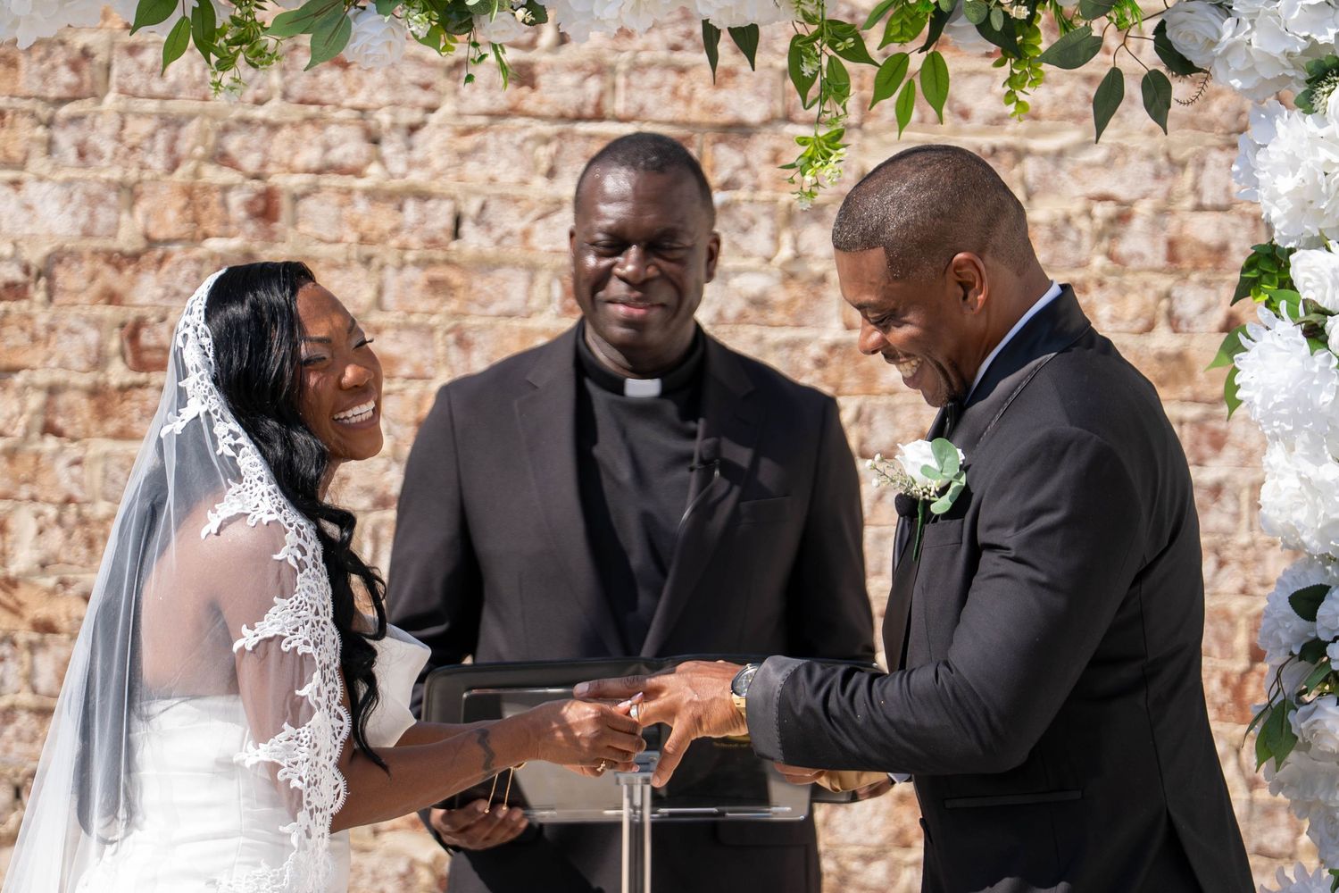Bride and groom exchange vows at outdoor wedding ceremony with officiant at acrylic podium, stone wall backdrop.