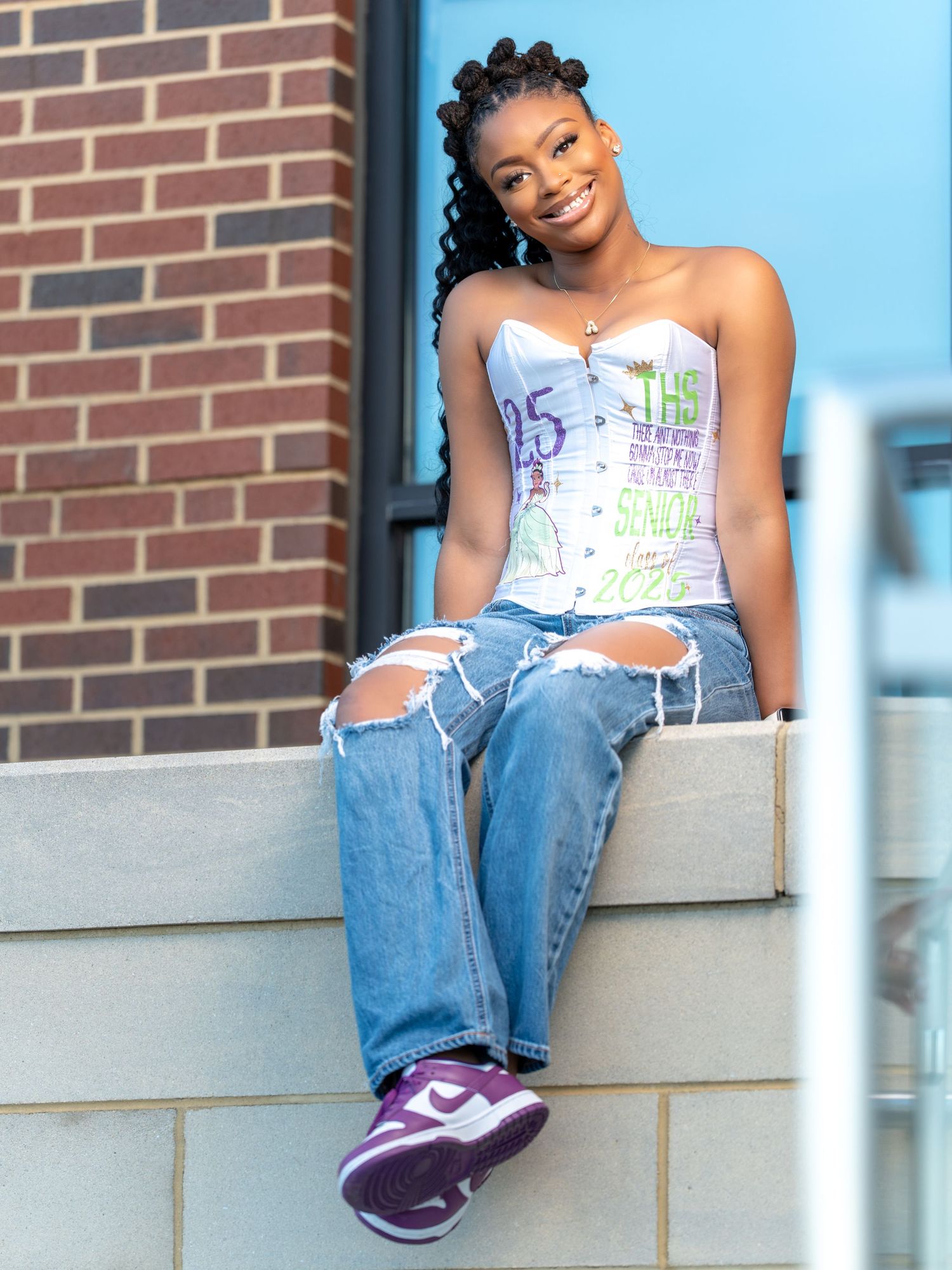 A person in a white patterned top and blue jeans sits casually on a concrete ledge against a brick wall.