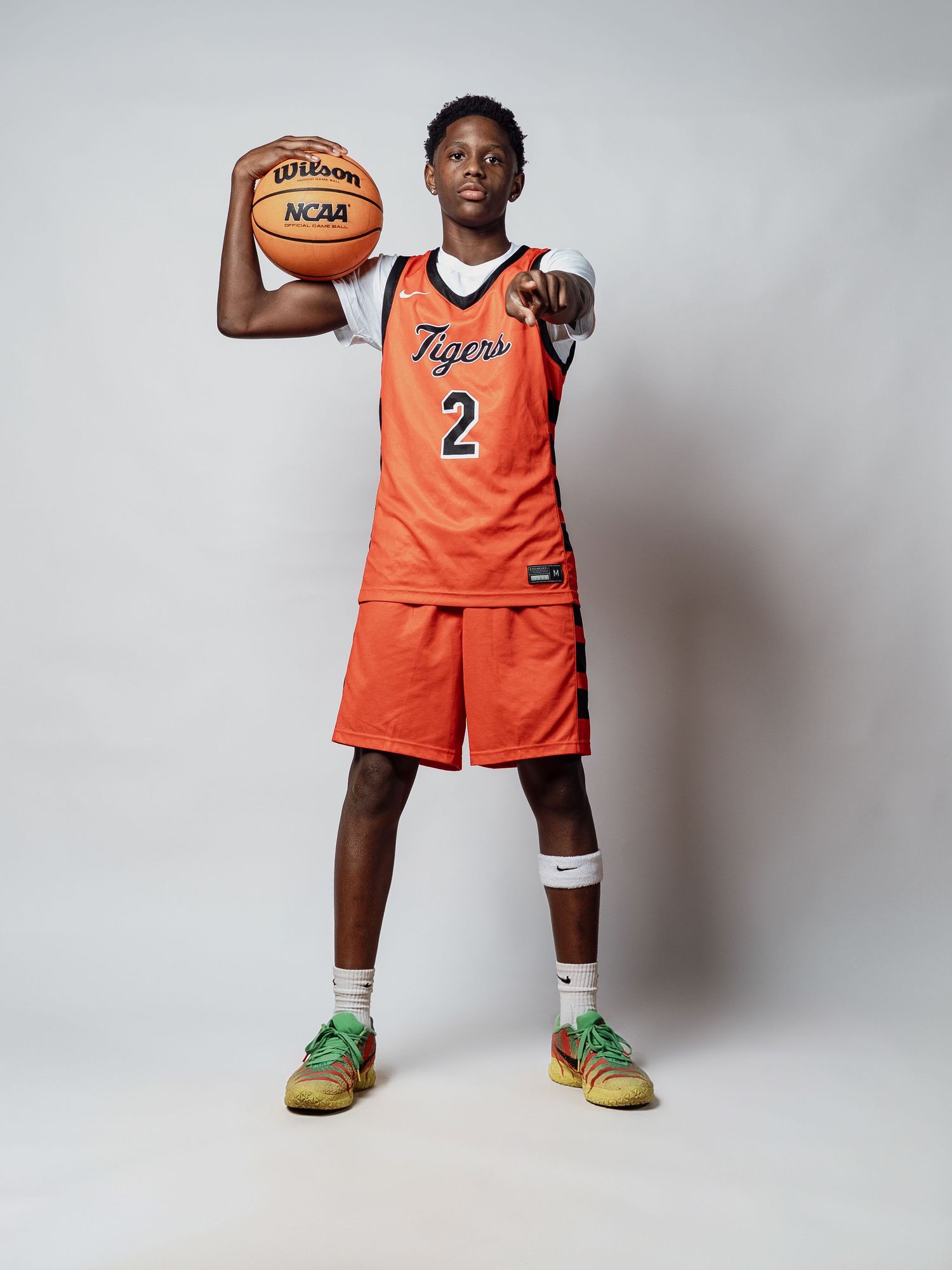 Young basketball player in an orange Tigers jersey number 2 holds a Wilson NCAA basketball and points at the camera in a studio portrait.
