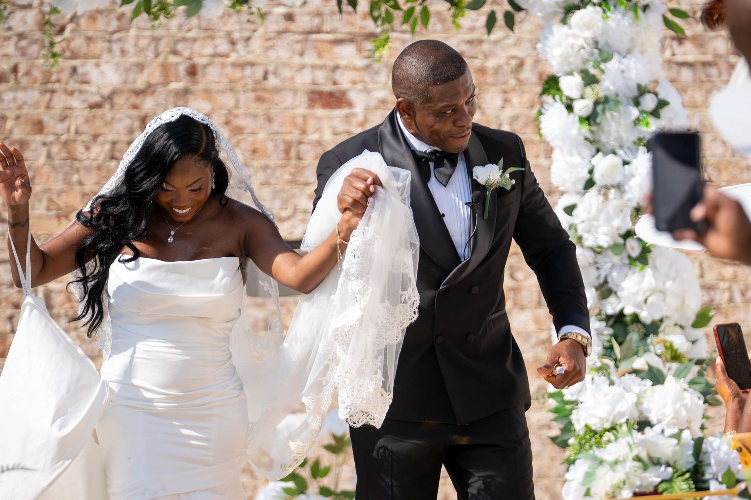 A joyful Black couple celebrates their wedding ceremony outdoors, bride in white gown and groom in black tuxedo.