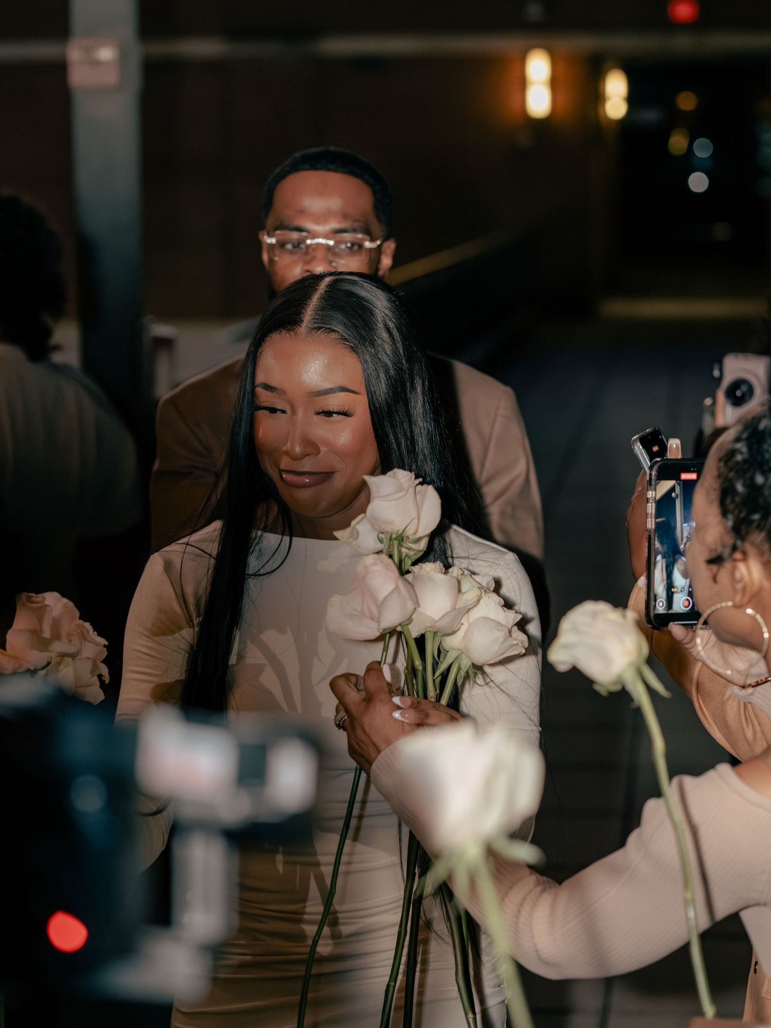 A woman in a beige dress holds white roses while being celebrated at an evening event with guests capturing the moment.