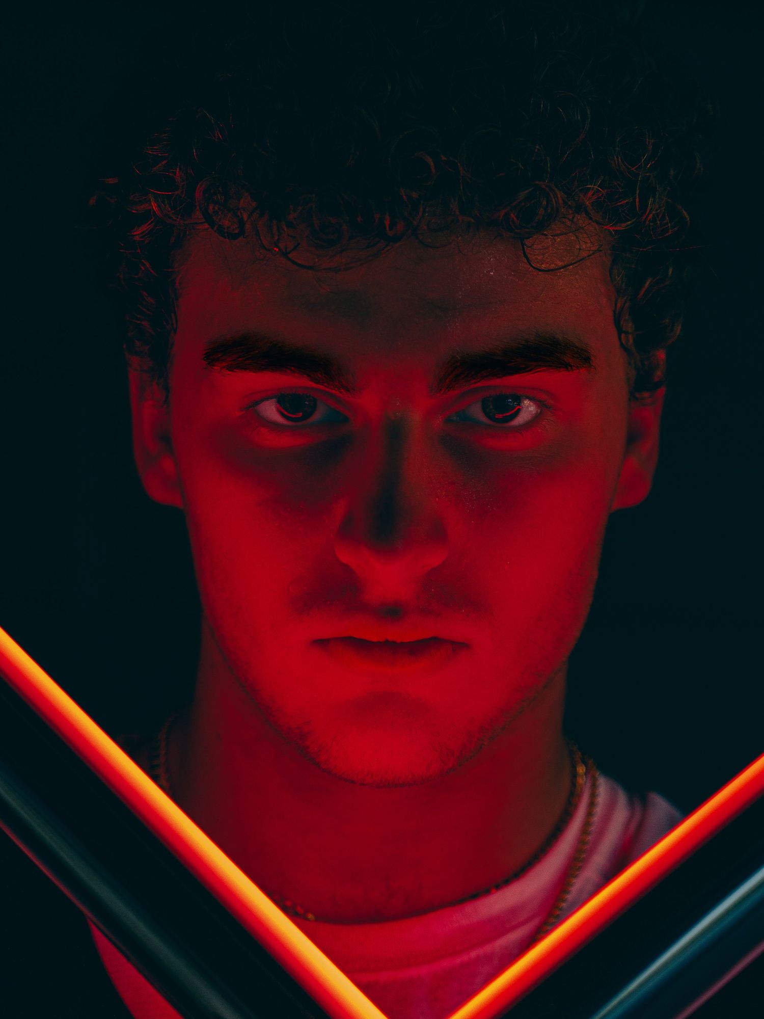 Close-up portrait of a curly-haired young man with an intense stare, lit in dramatic red light between two glowing neon tubes.