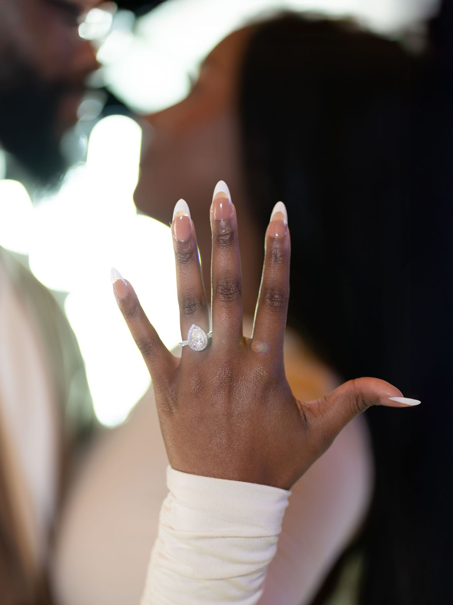 Close-up of a woman displaying a large diamond engagement ring with long white almond-shaped nails while embracing her partner.