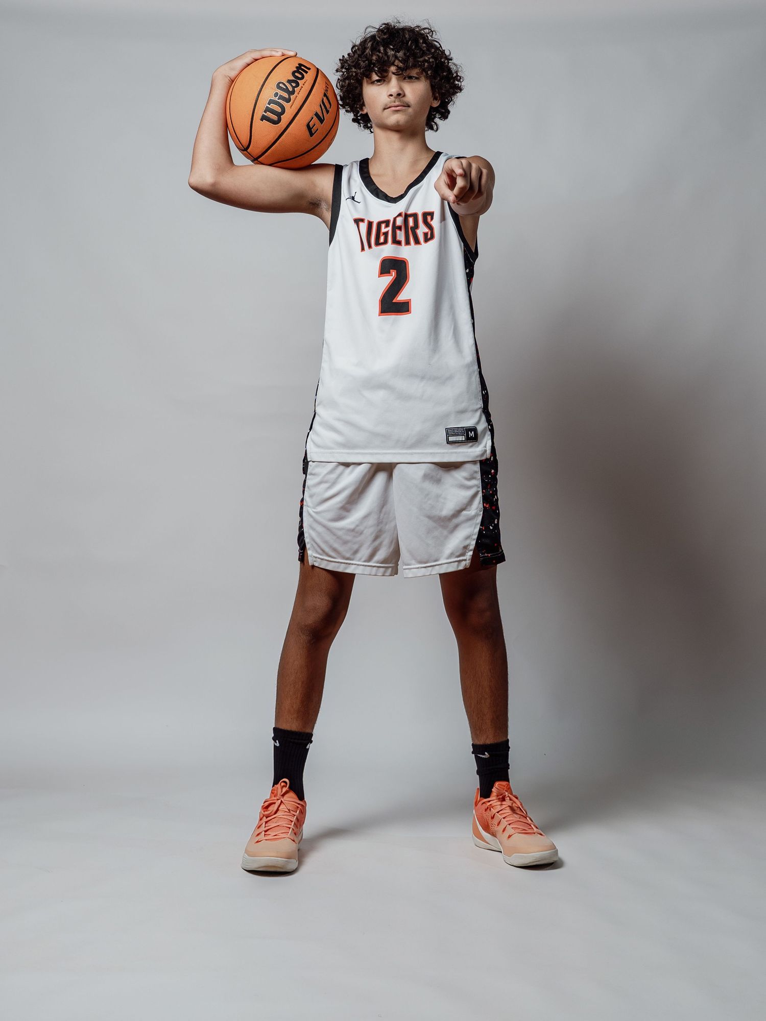 Young basketball player in a white Tigers number 2 jersey holds a Wilson basketball and points at the camera against a gray studio backdrop.
