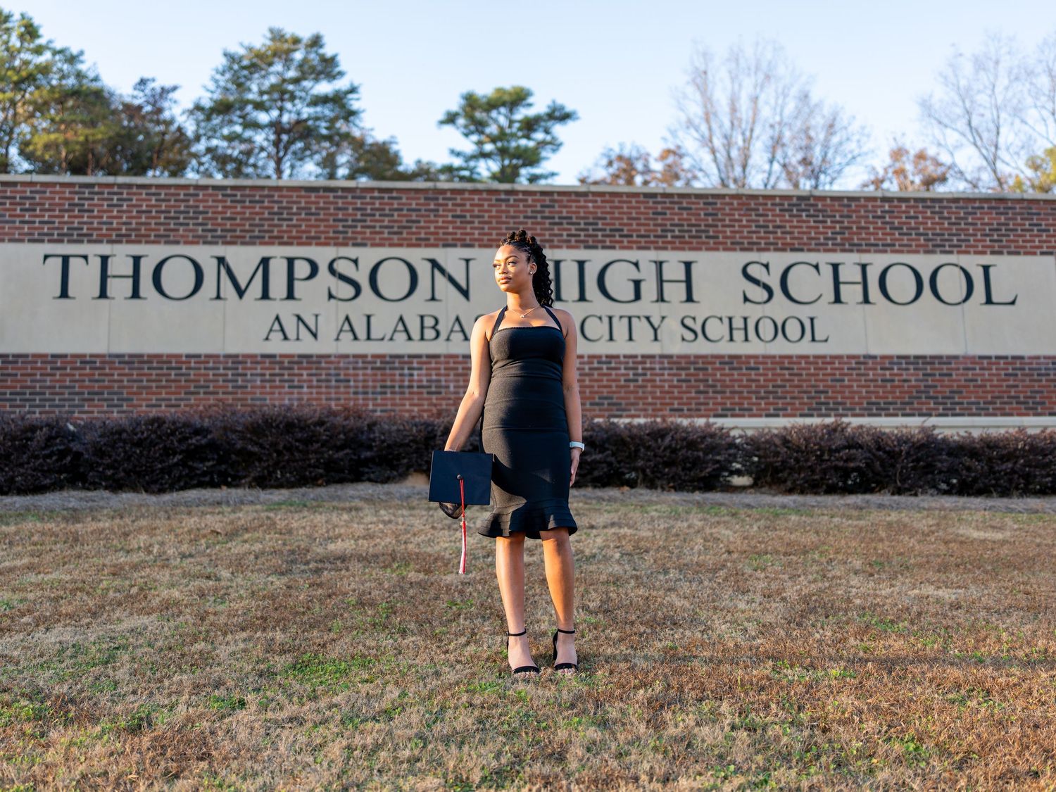 Student stands in front of Thompson High School sign on brick wall during graduation photo session.