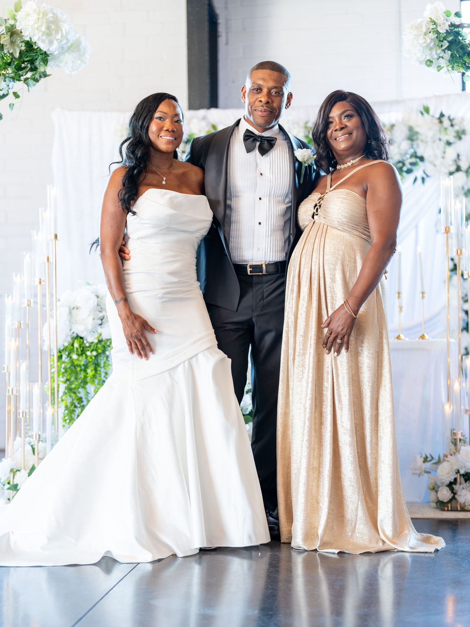 Bride in white strapless gown poses with groom in tuxedo and guest in gold dress at elegant wedding venue.