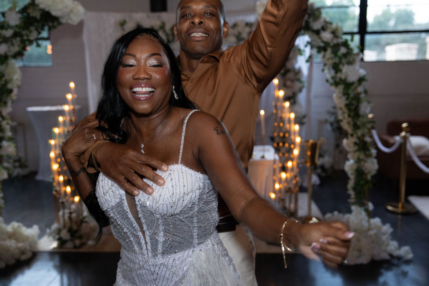 A joyful couple dances at their wedding reception, bride in beaded gown, groom in brown shirt, with floral decor behind.
