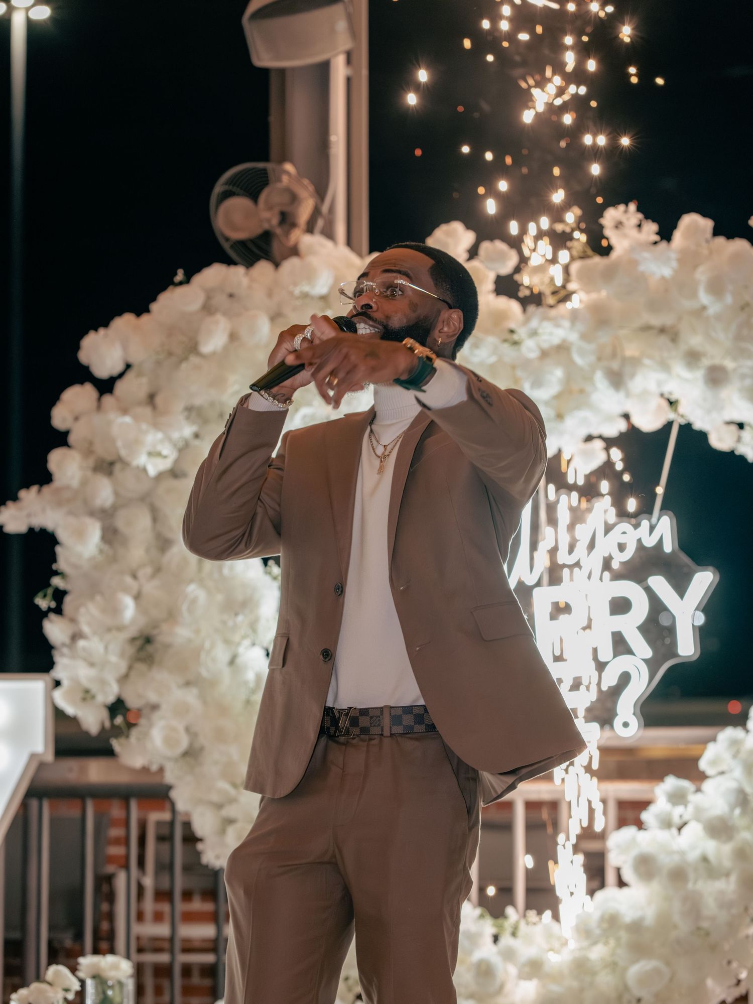 Man in tan suit singing into microphone at romantic proposal setup with white flowers and Will You Marry Me neon sign.