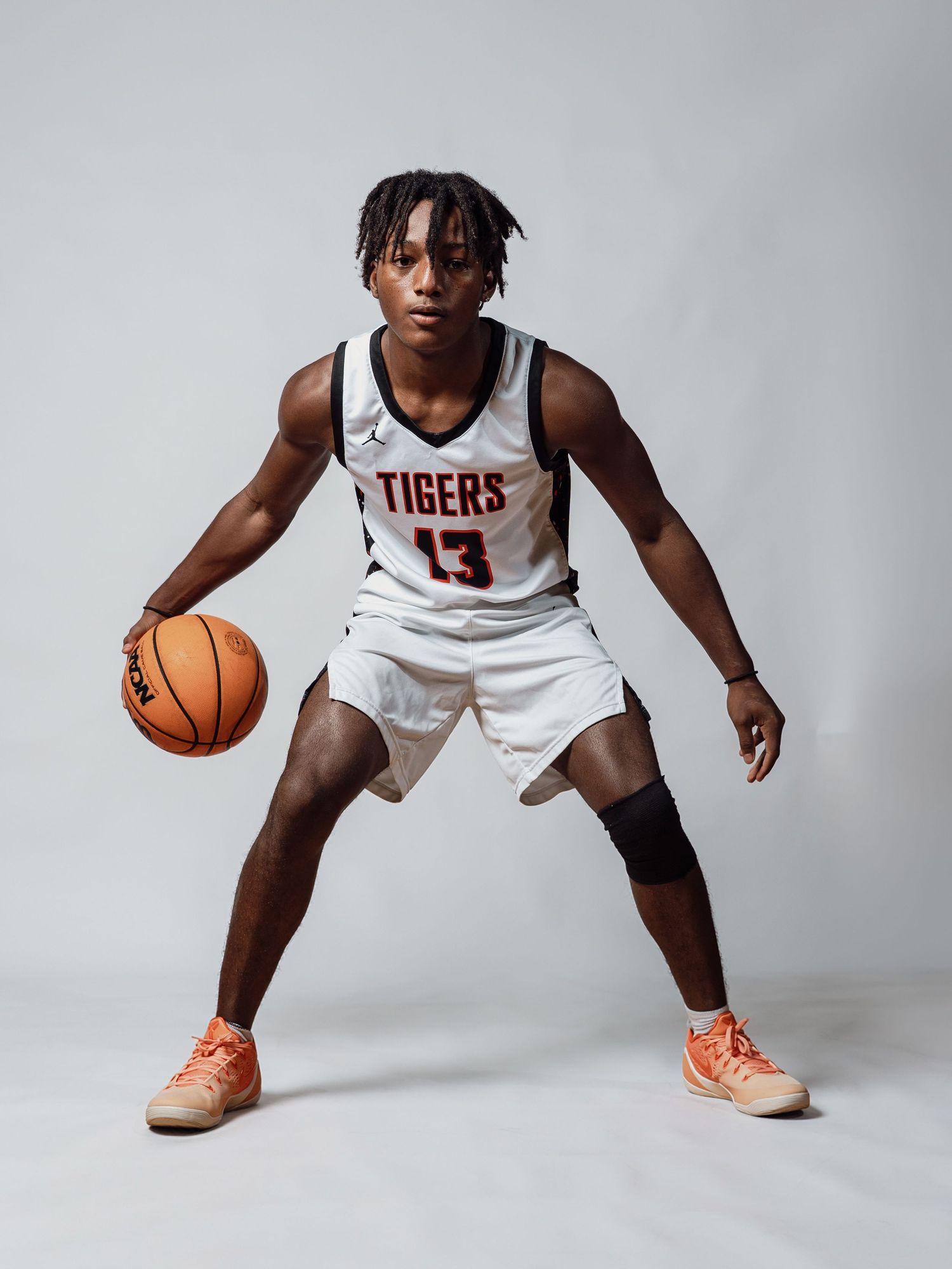 Basketball player in a white Tigers number 13 jersey crouches in a dribbling stance while holding an orange basketball in a studio.