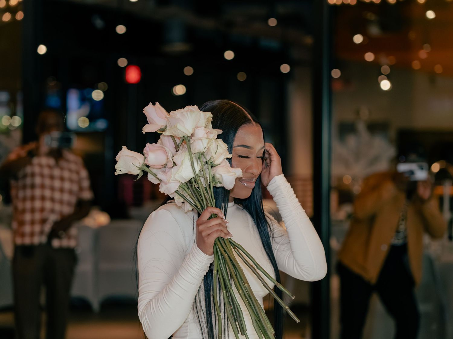 Person in white sweater holding a bouquet of pink flowers while talking on phone in dimly lit indoor setting.