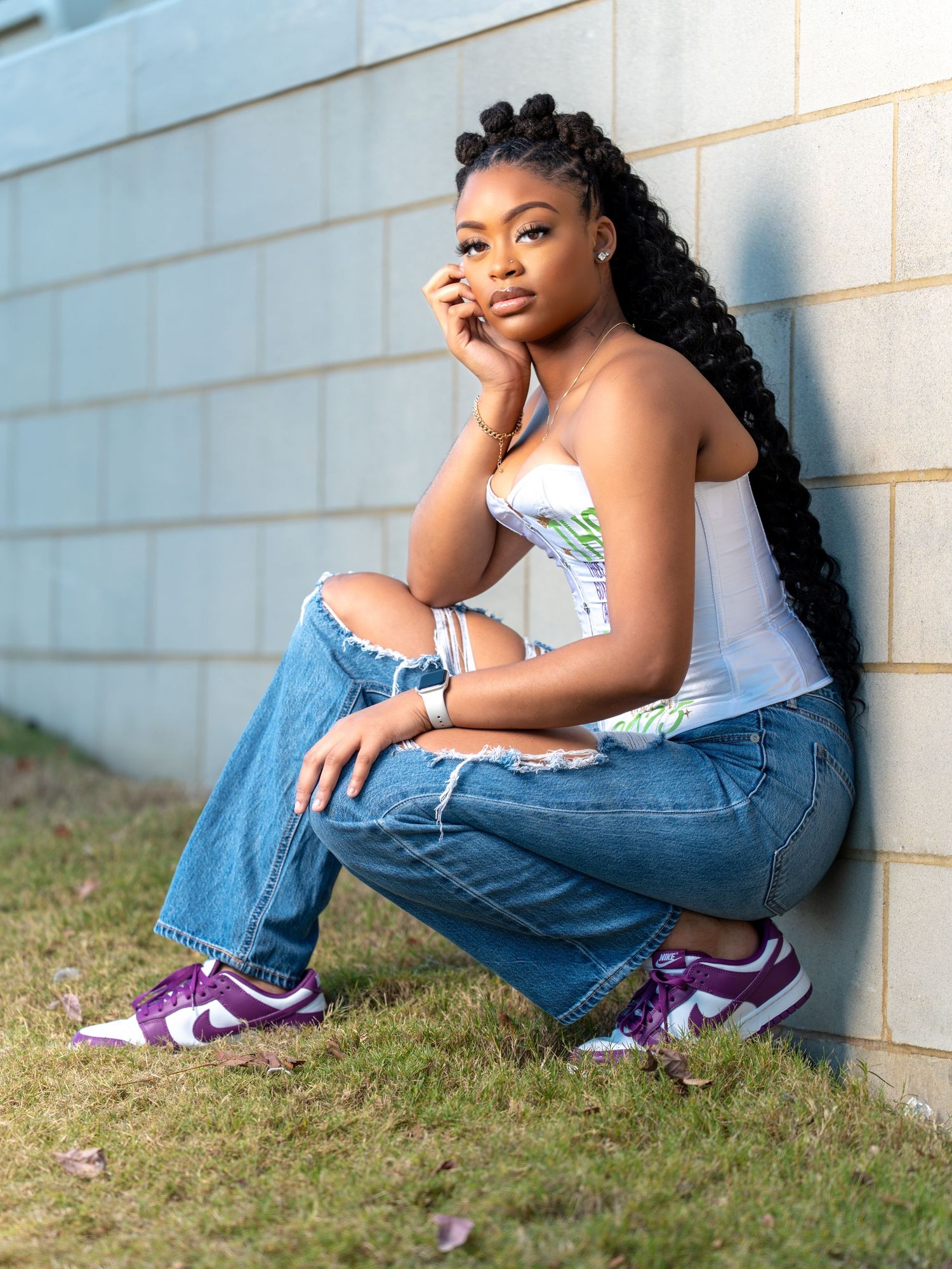 A person in casual attire sits against a wall wearing purple Nike sneakers and ripped jeans.