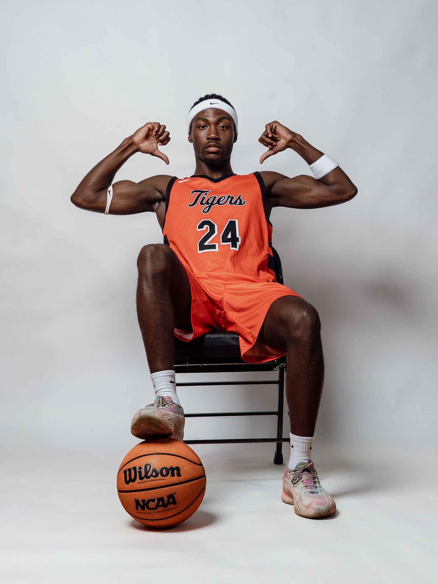 Basketball player in an orange Tigers number 24 jersey sits on a chair, flexing both arms with one foot on a Wilson NCAA basketball.