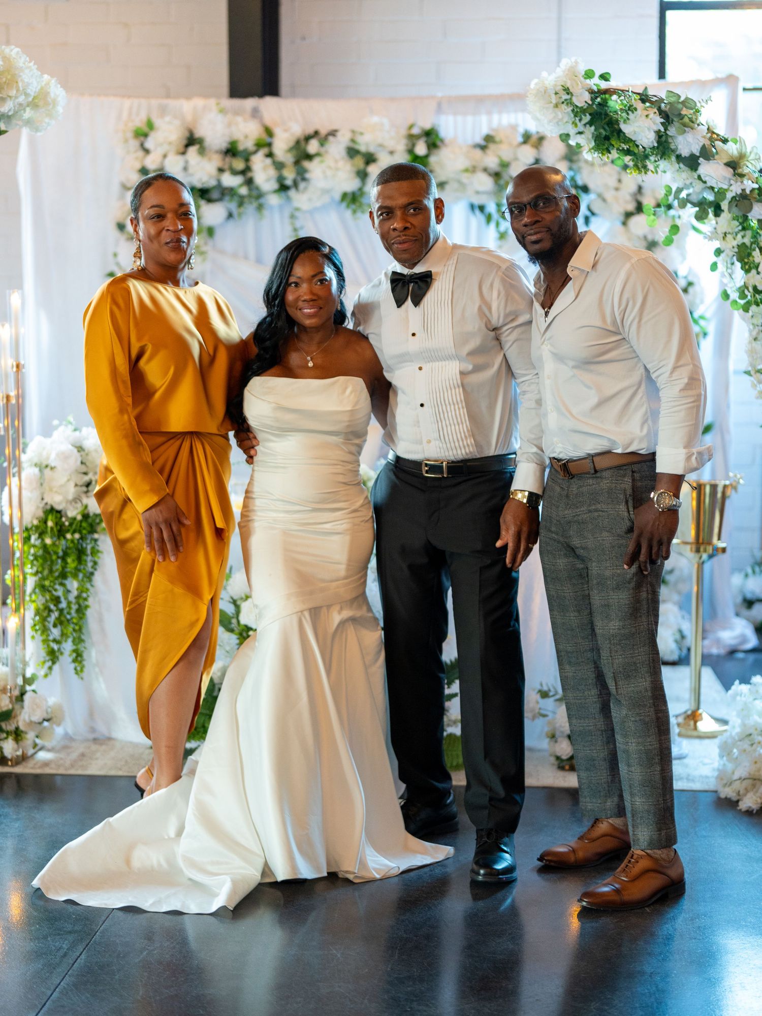 A bride in a white gown poses with groom in tuxedo and two guests at an elegant floral wedding backdrop.