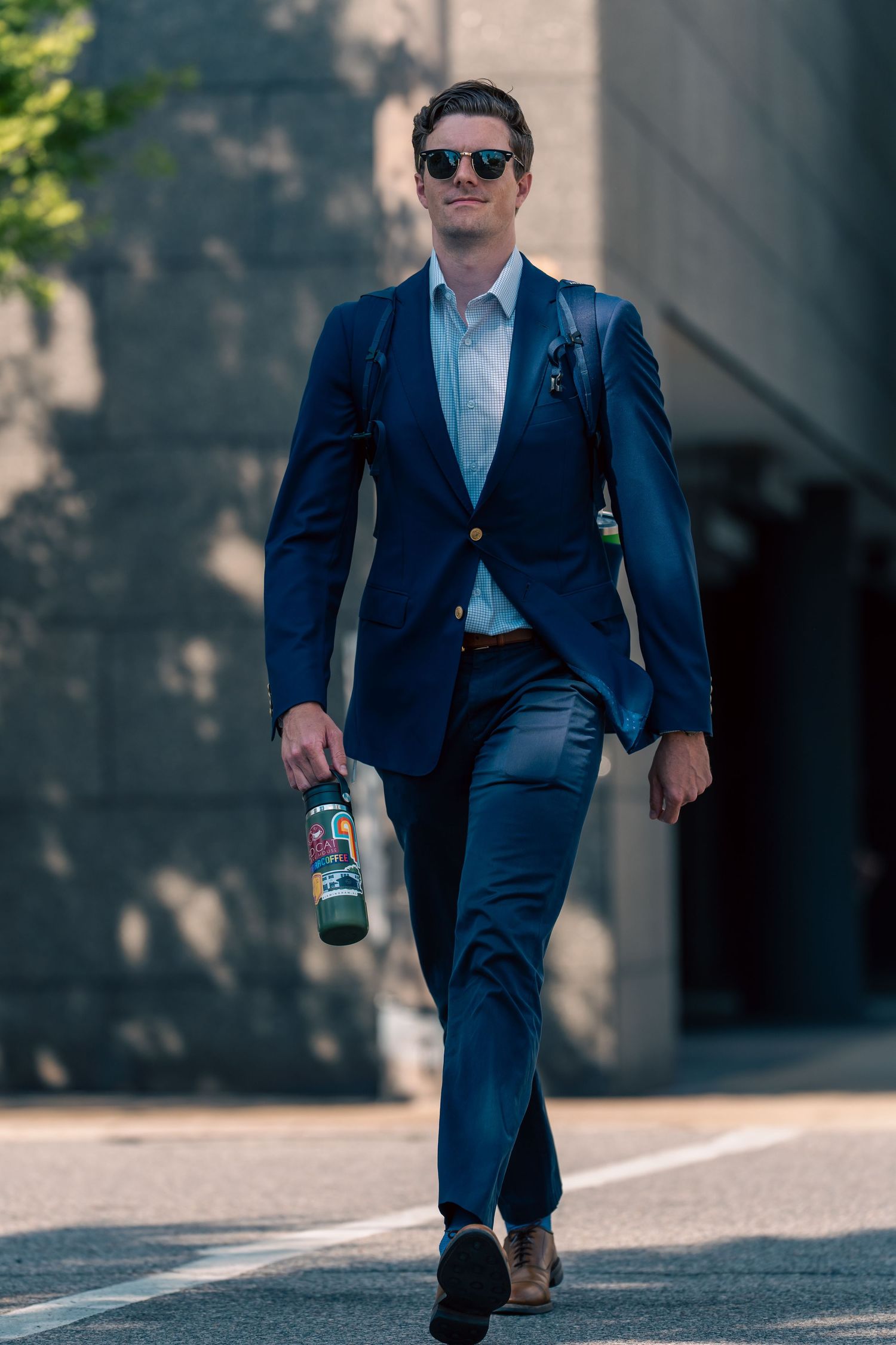Stylish person walking in navy blue suit and sunglasses while carrying a drink bottle.