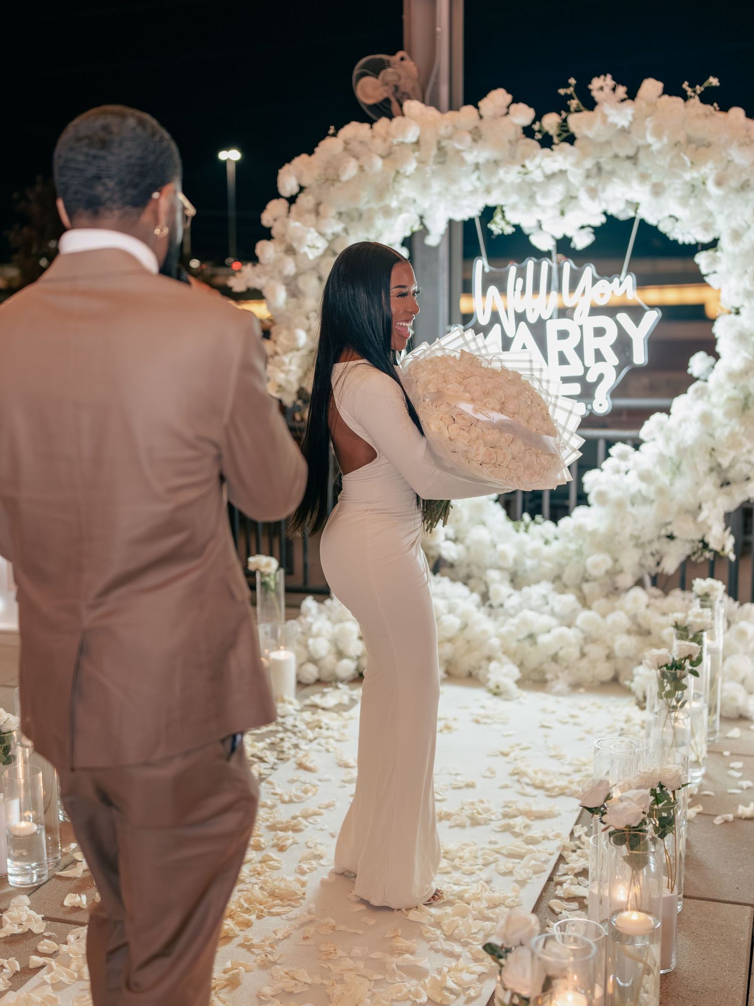 Romantic outdoor proposal setup with white floral heart arch, neon marry me sign, candles, and couple in elegant attire at night.
