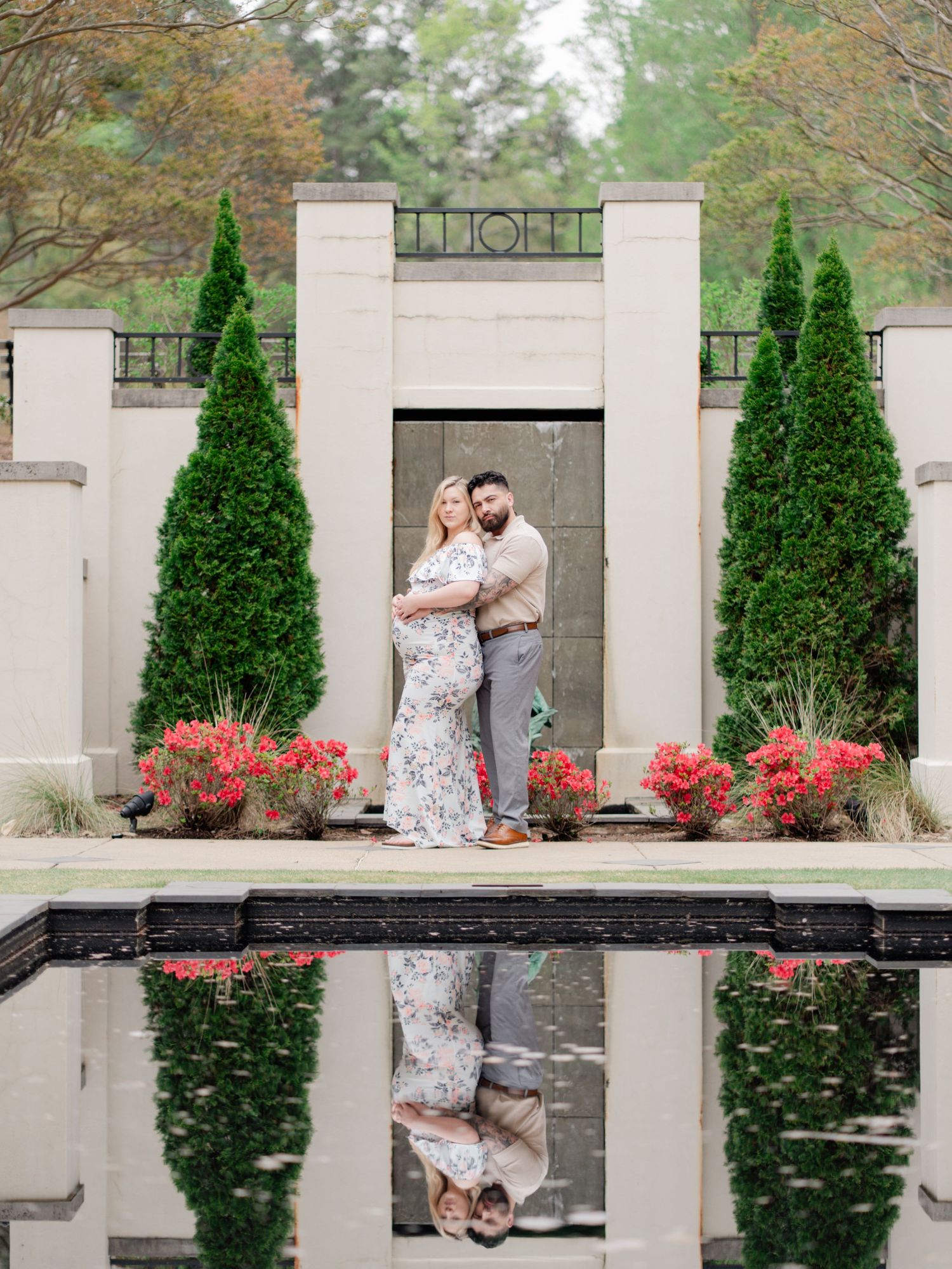 Couple embracing in front of a stone archway with evergreen trees and red azaleas, their reflection visible in a still garden pool.