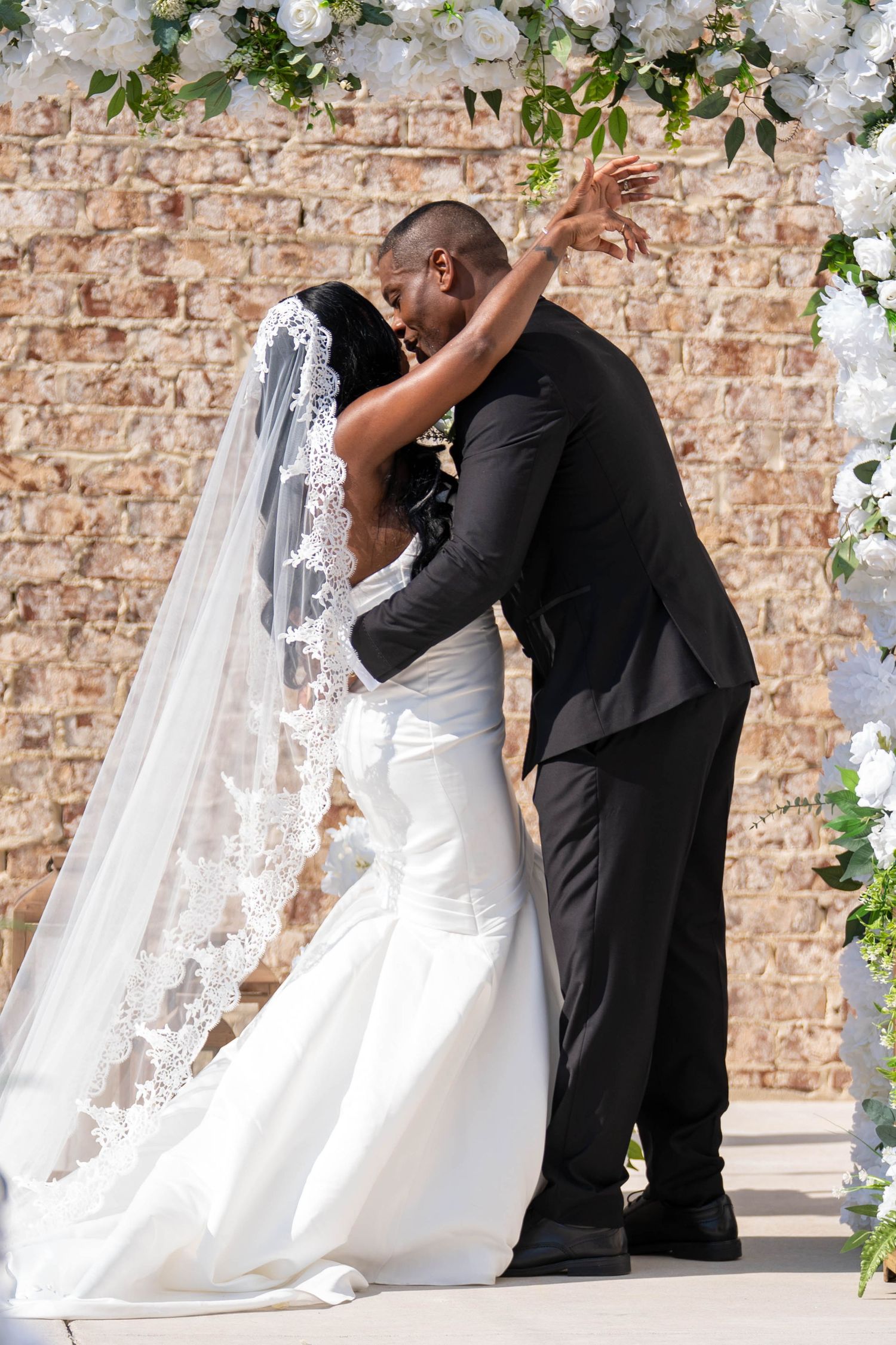 Bride and groom share first kiss under floral arch, bride wearing lace veil and white gown, groom in black suit.
