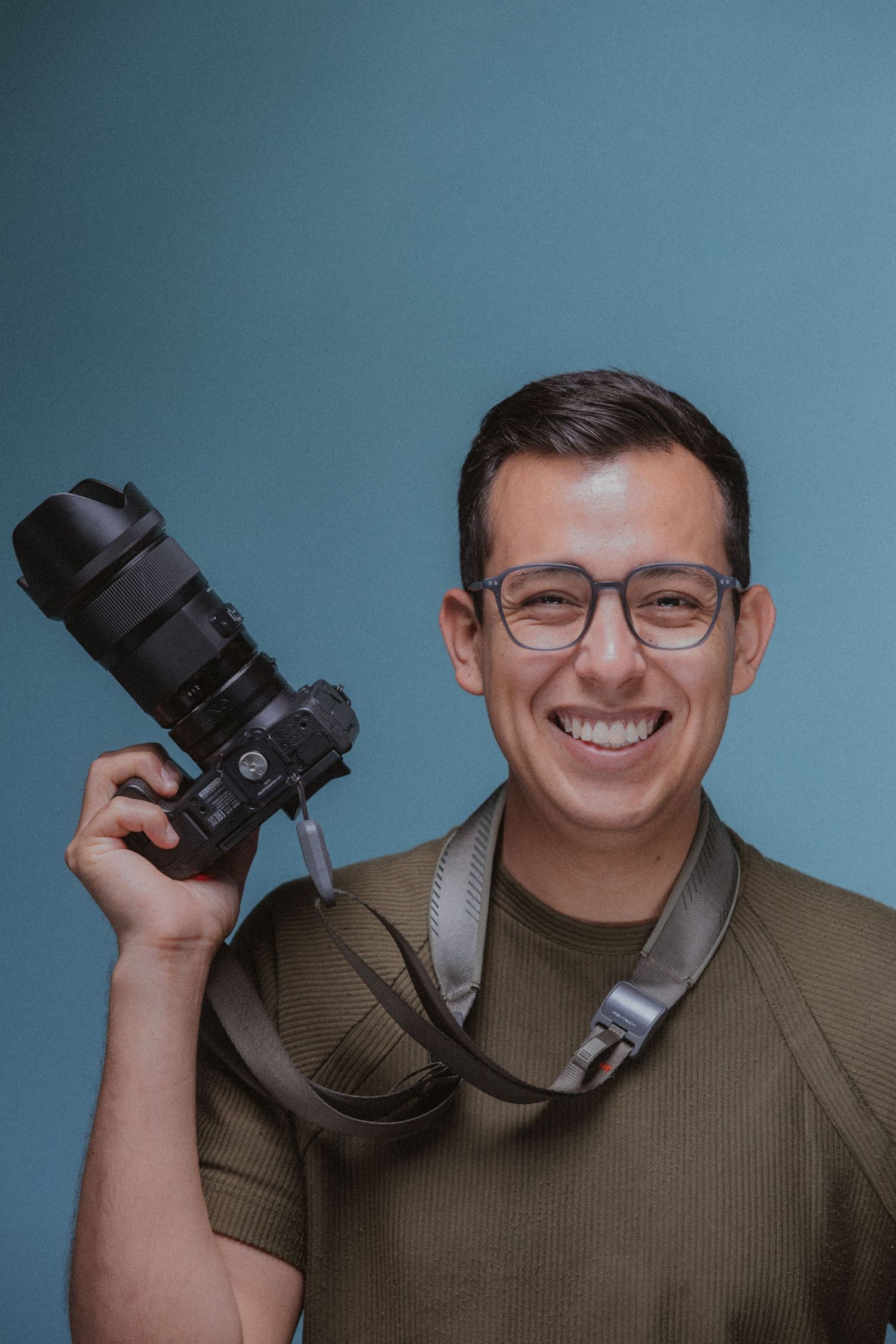 Smiling photographer wearing glasses demonstrates camera poses against blue studio background.