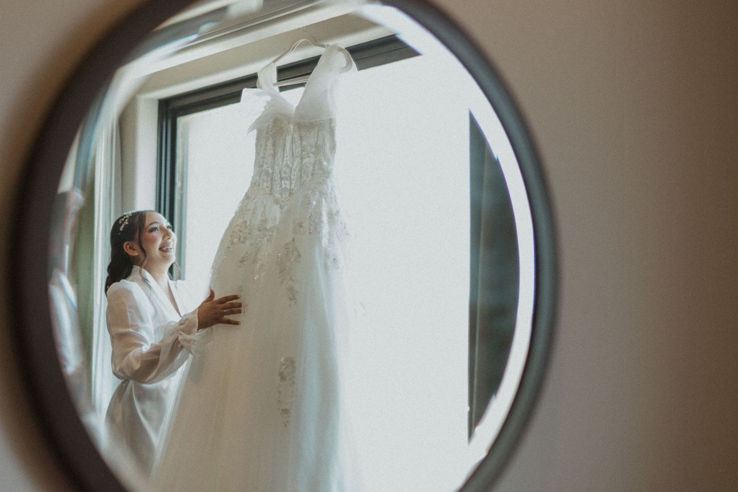 Multiple reflections in an oval mirror show a wedding dress by a window.