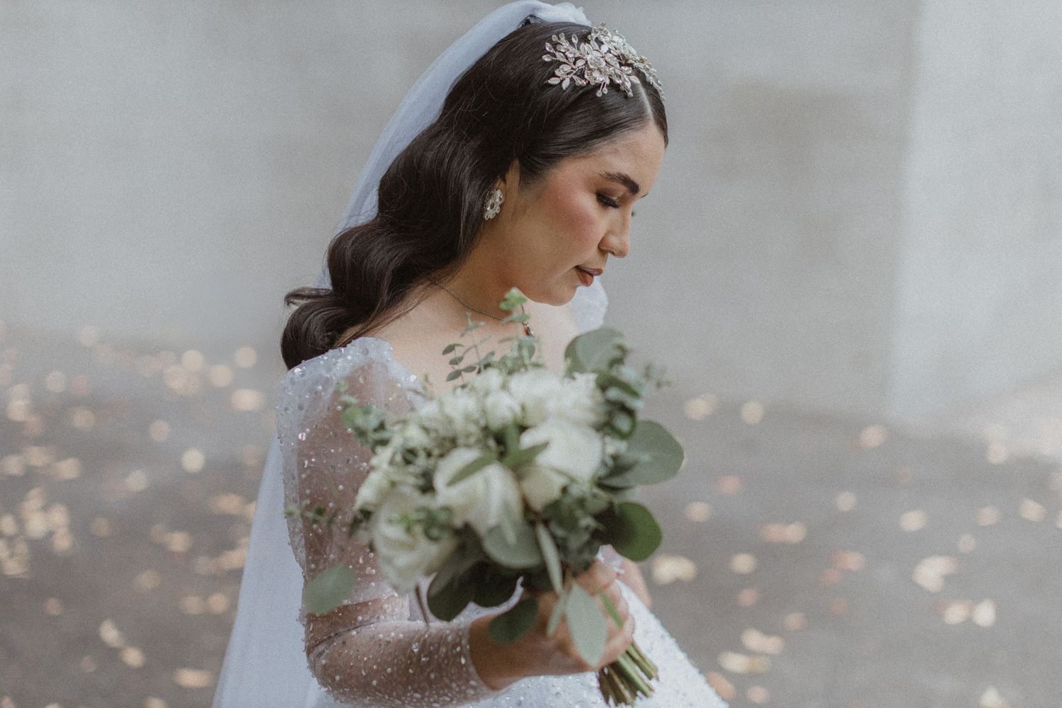 Bride in lace gown holds white floral bouquet against bokeh background.