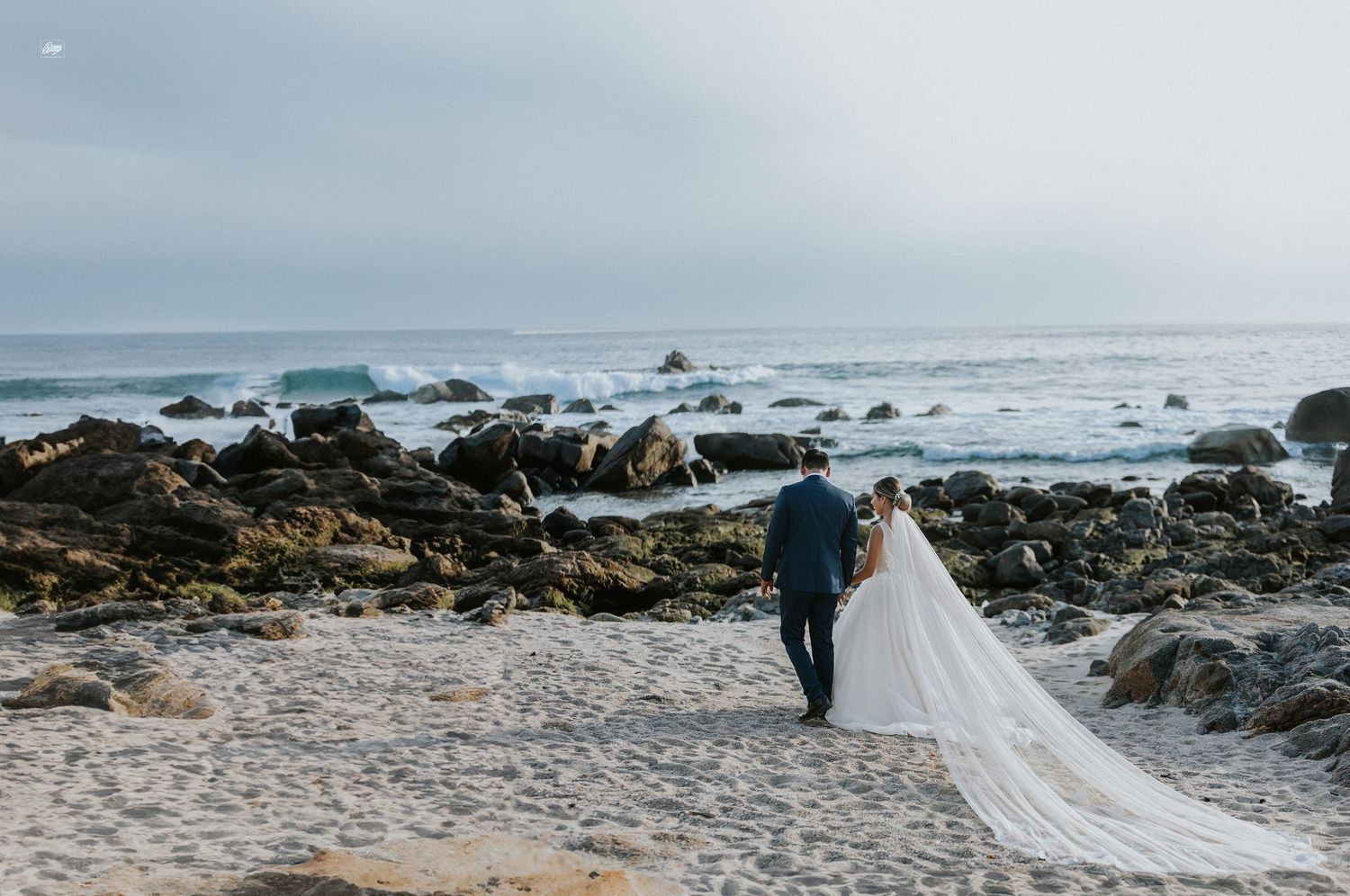Couple walking on rocky beach with long wedding dress train trailing behind.