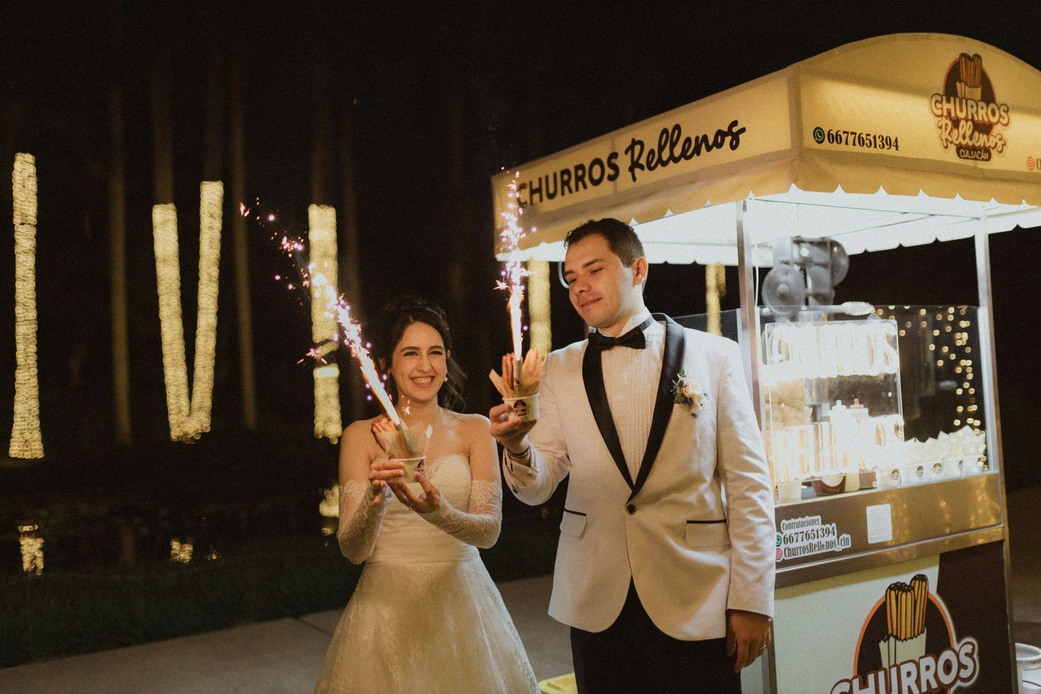 A wedding couple poses near a snack vendor cart at night with decorative lights.