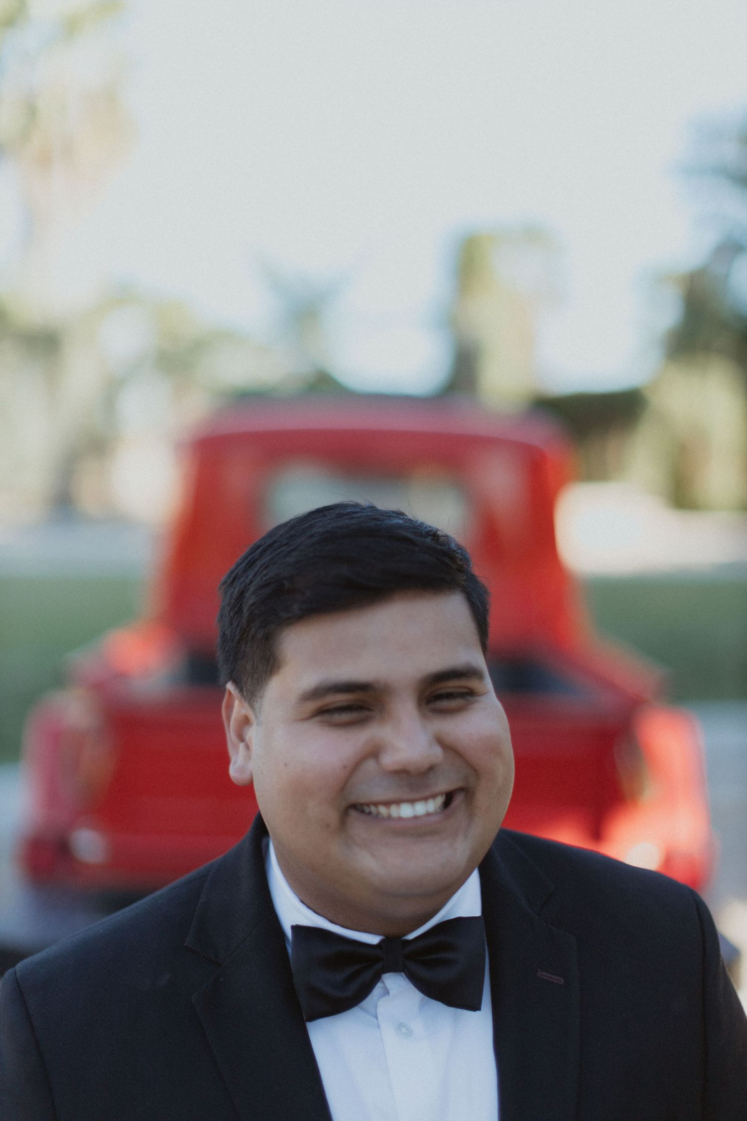 Groom in classic black tuxedo and bowtie smiling with red vintage car in background.