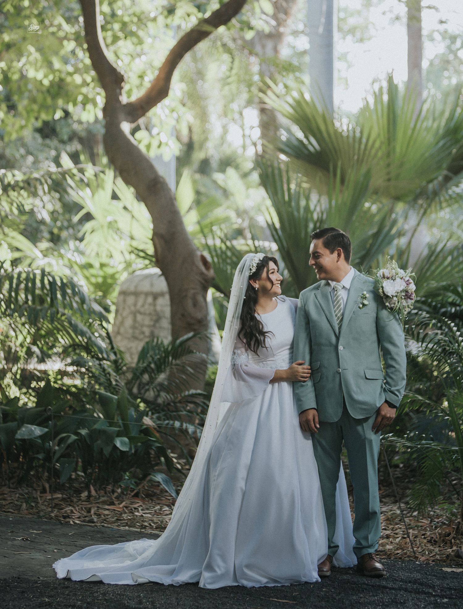 A couple in wedding attire standing together in a lush garden setting with palm trees.