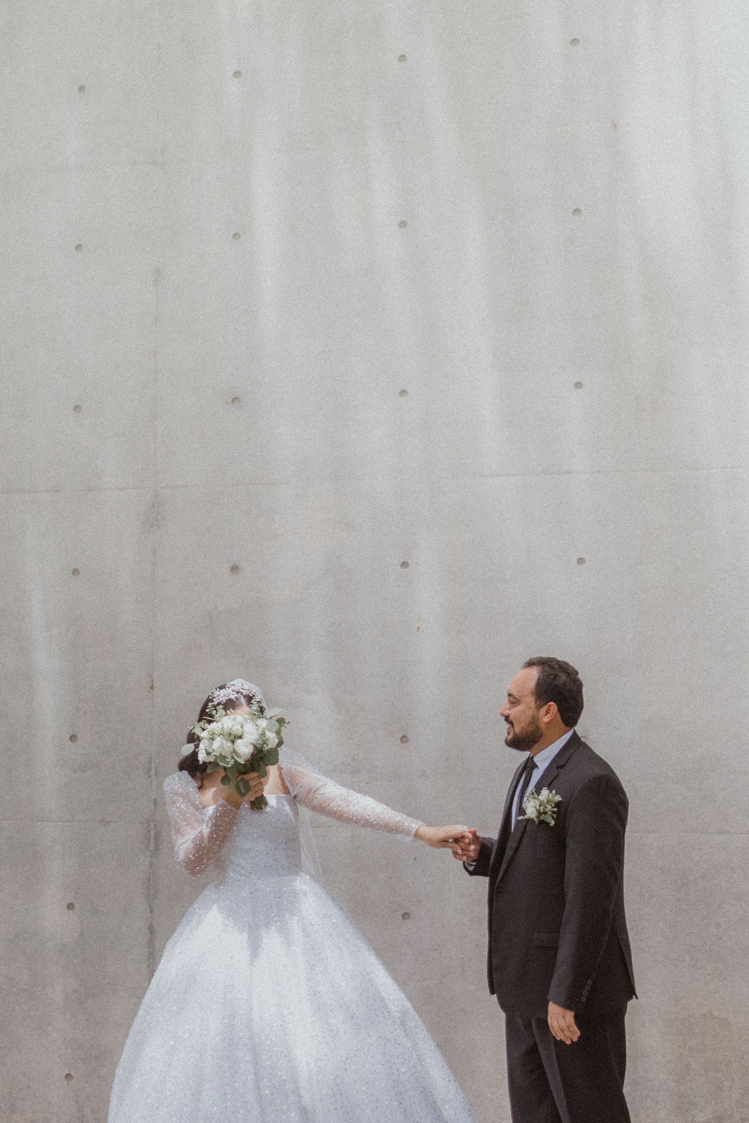 A bride in a white gown and groom in a dark suit share a playful moment against a concrete wall.