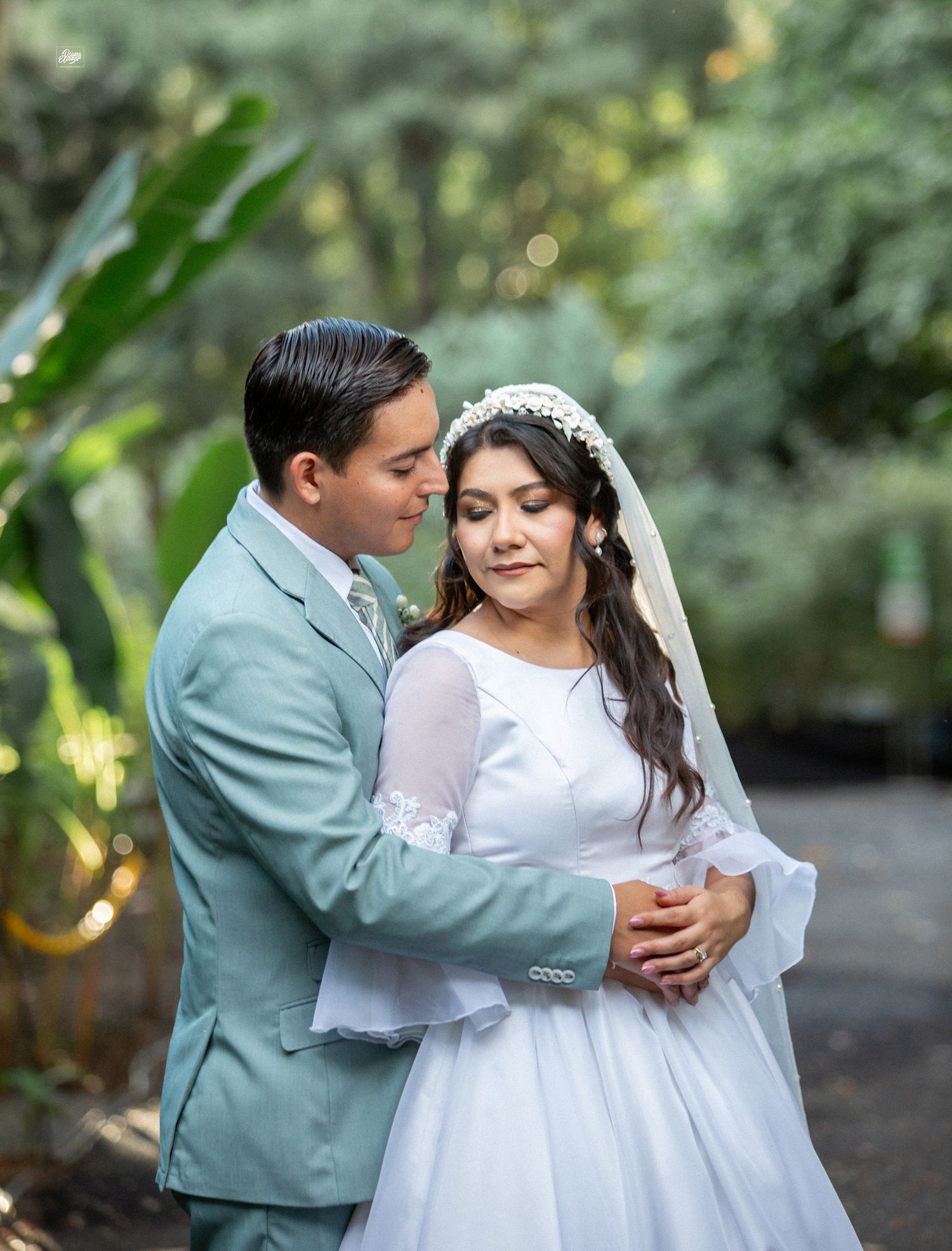 A couple in wedding attire embracing in a garden setting with tropical foliage.
