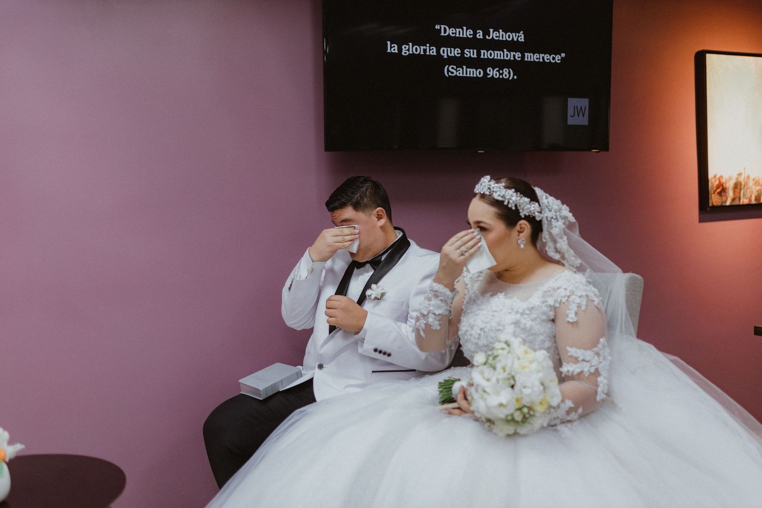 A couple in wedding attire sits in front of display screen during ceremony.