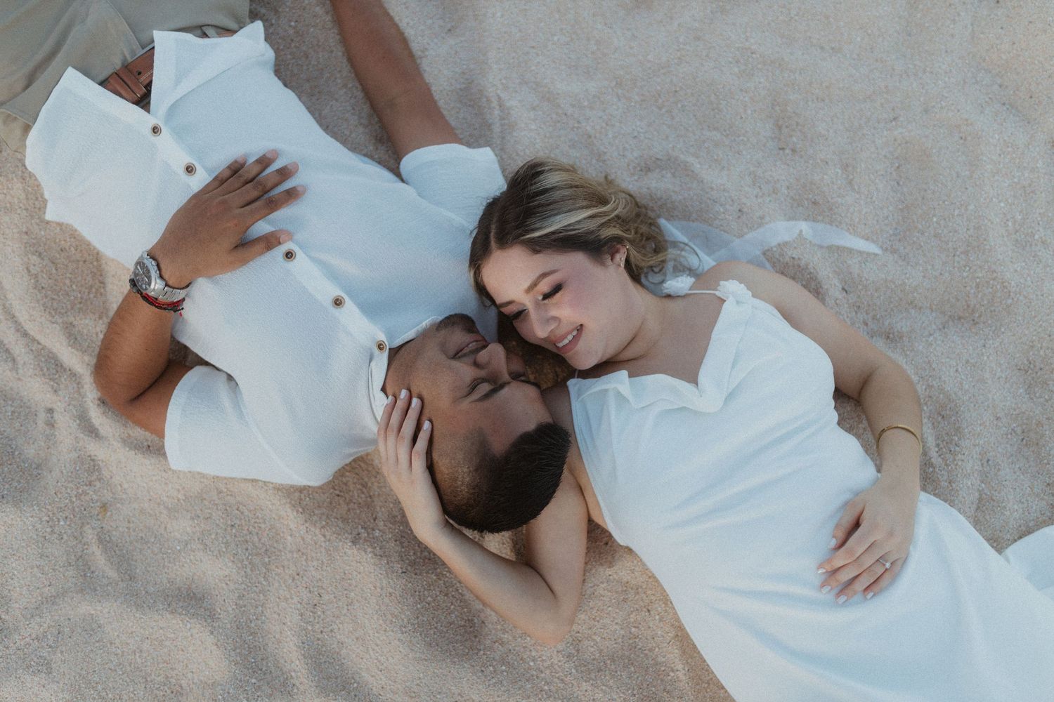 Couple in white clothing laying together on sandy beach during romantic photoshoot.