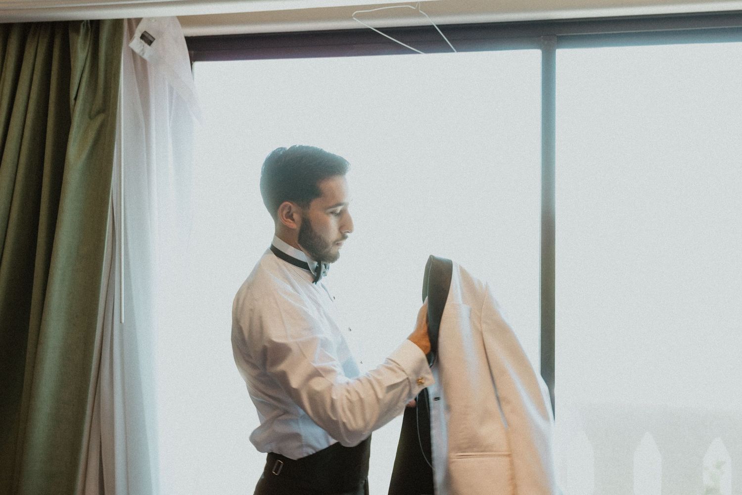 Person in formal attire preparing by adjusting their jacket sleeve near a window.