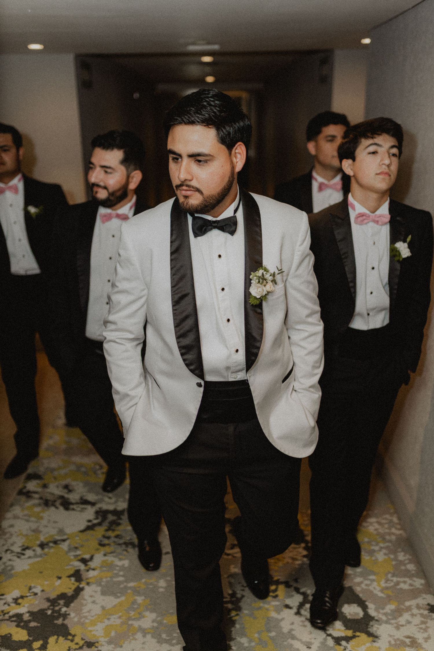 Groomsmen in black and white tuxedos walk through a hallway with confetti on the floor.