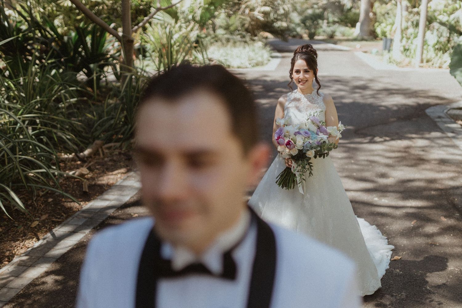 Elegant wedding party poses in dappled sunlight under garden trees.