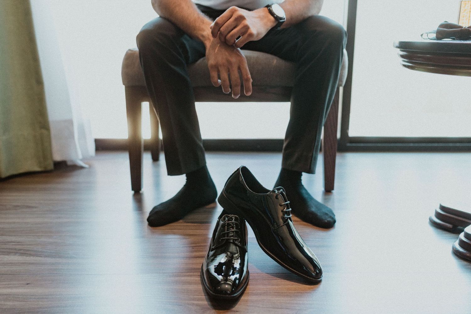 Black dress shoes placed on hardwood floor next to someone sitting in a chair.