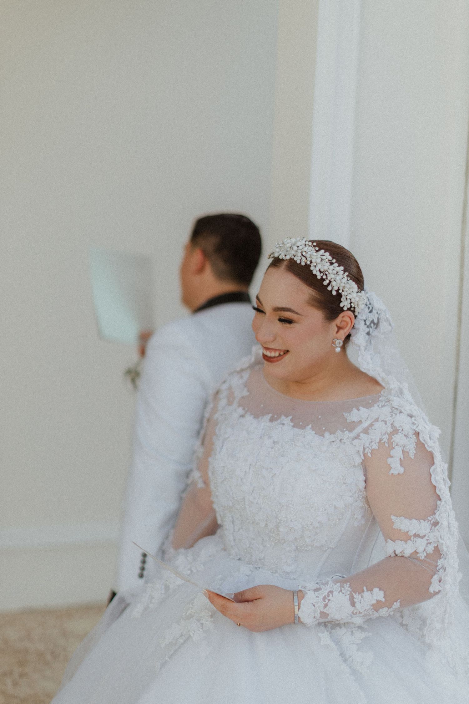 A bride in a lace ballgown and tiara smiles while reading during her wedding ceremony.
