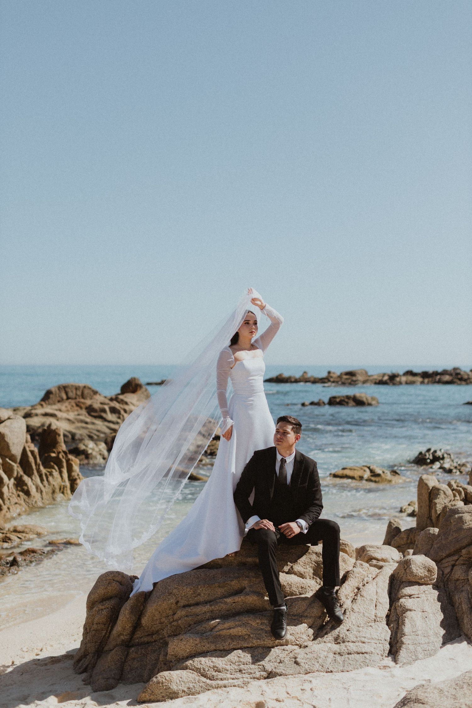 A flowing wedding dress veil blows in the wind on a rocky beach coastline.